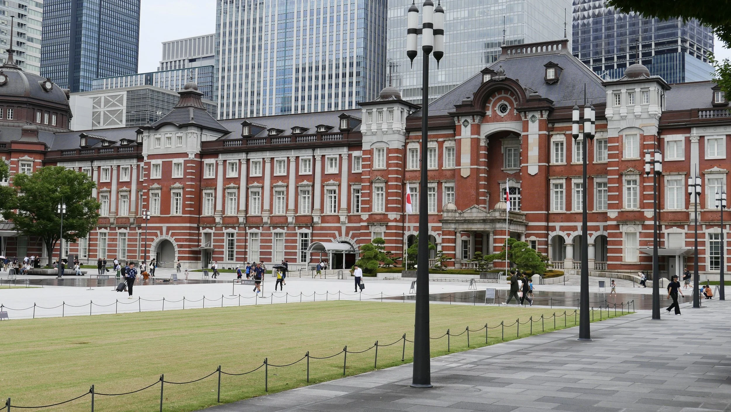 A historic red-brick building with white accents and a gray roof, surrounded by a large open plaza with people walking and sitting, located in an urban area with tall modern skyscrapers in the background.
