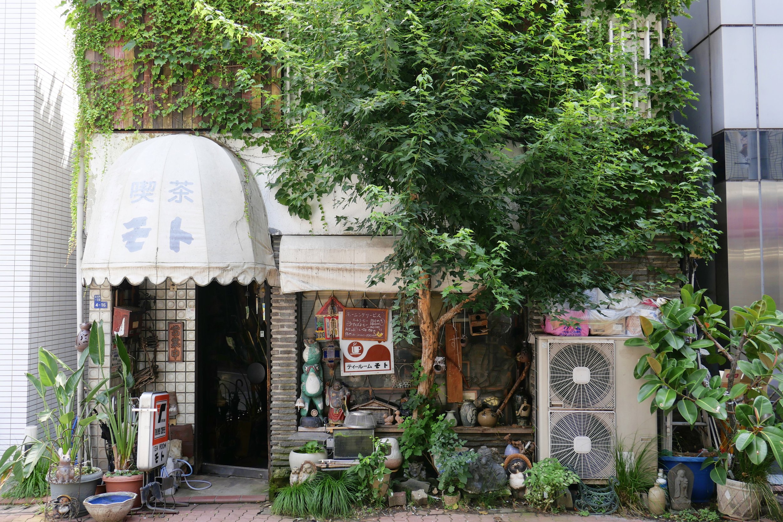 A storefront with an abundance of green plants and vines covering the upper facade, partial signage with Japanese characters, and various decorative items and plants displayed at the entrance, including large air conditioning units on the right, and 