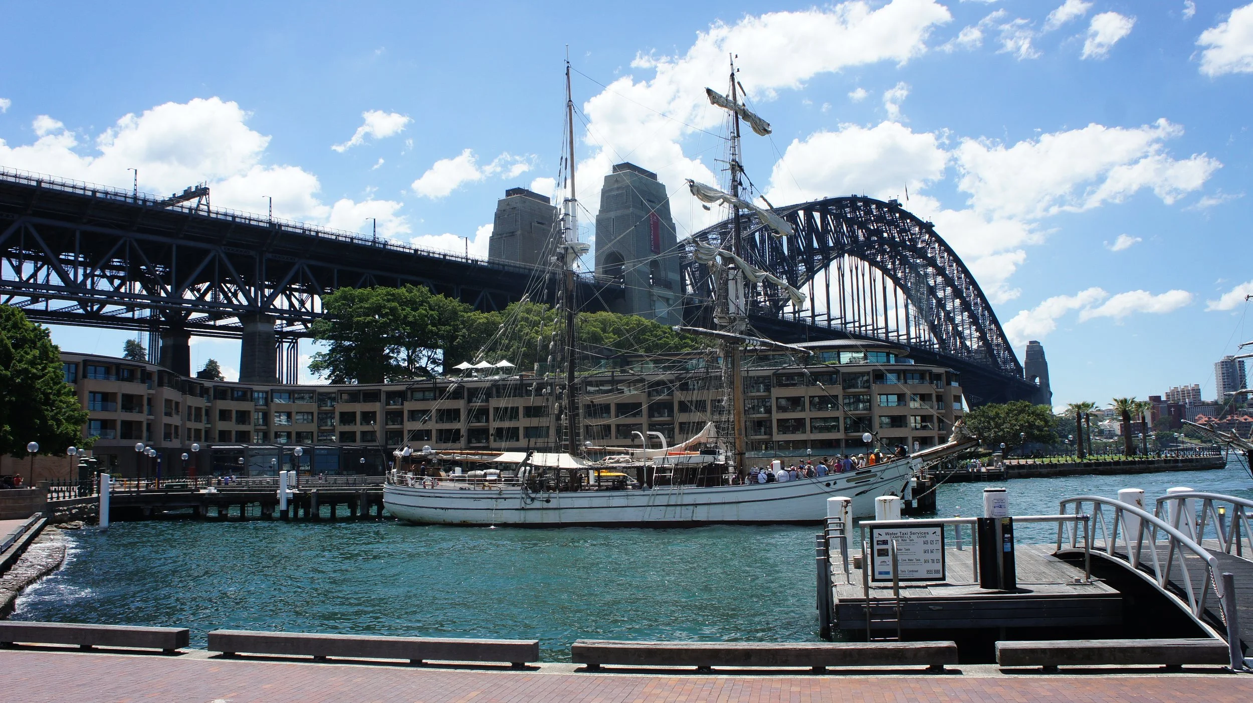 A harbor scene with a sailboat docked in the foreground, tall buildings, including a bridge, and a partly cloudy sky in the background.