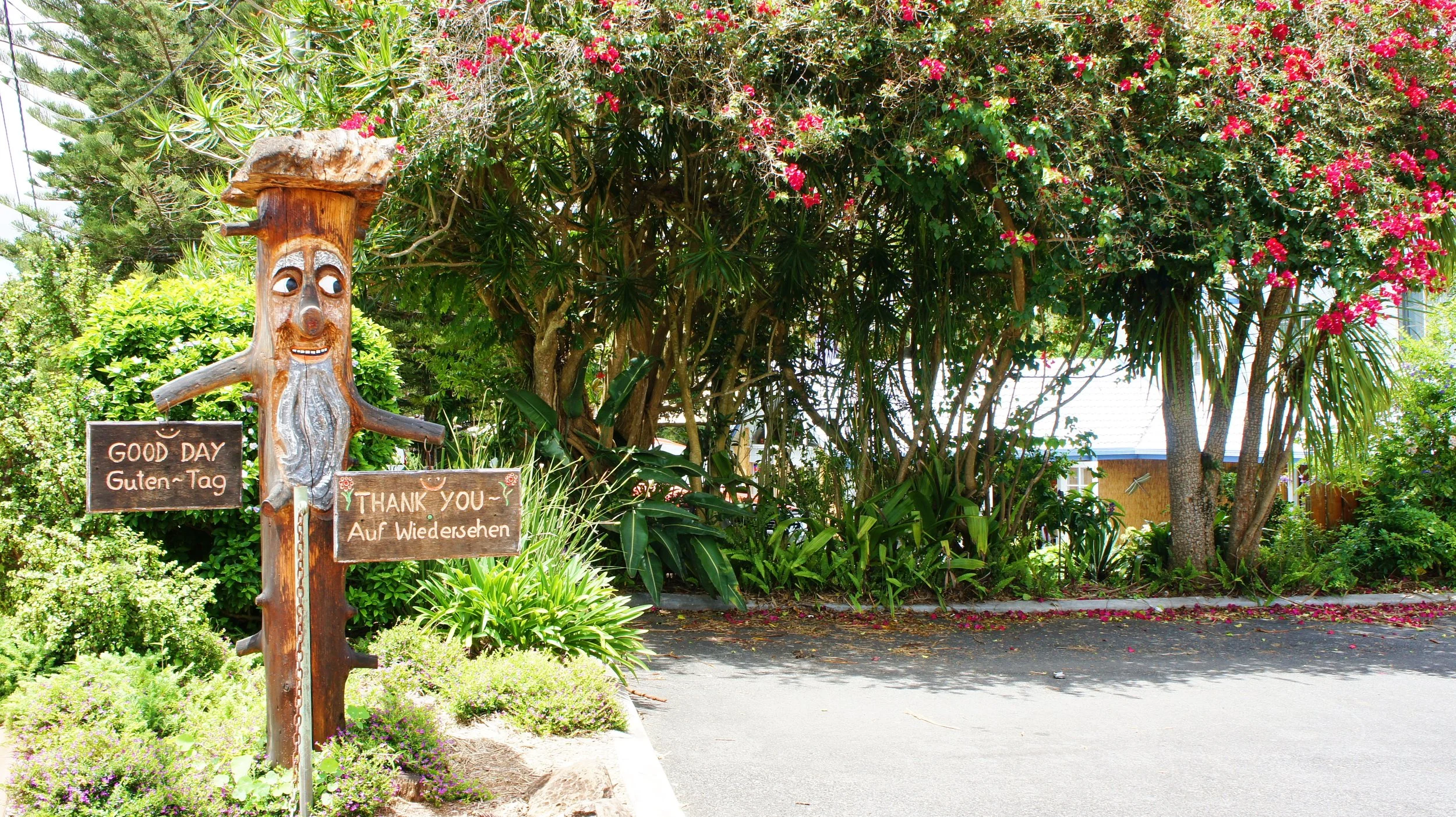 A wooden smiling tree sculpture with eyes, nose, and mouth in a garden surrounded by green plants and pink flowers. Two signs hang from it, one says 'Good Day Guten-Tag' and the other says 'Thank You Auf Wiedersehen'.