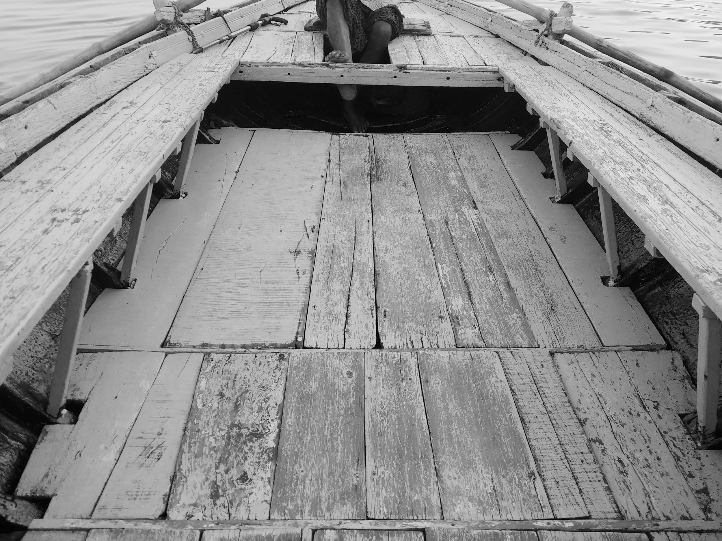 A black and white photo of a person sitting on a wooden dock by a body of water. The person appears to be reaching into or examining a hole beneath the dock. The dock is made of weathered wooden planks with some rough textures, and there are ropes an