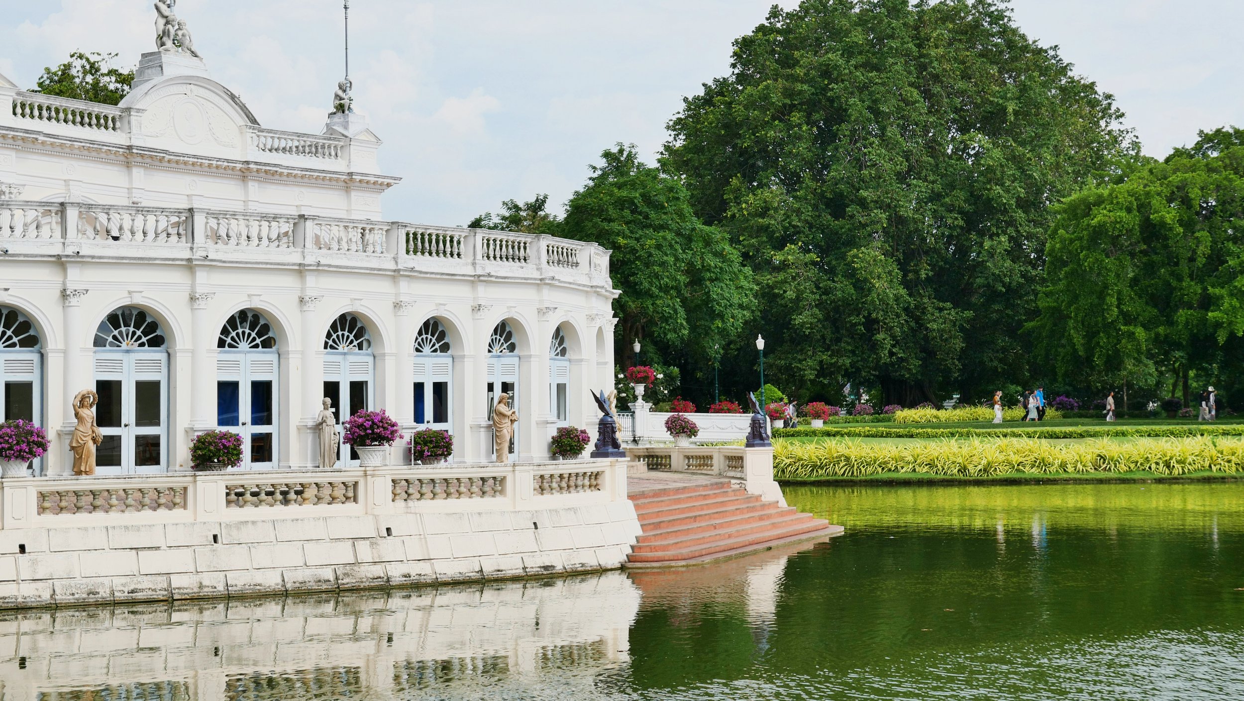 White ornate building with arches, sculptures, and potted plants along a river, surrounded by green trees and a park with people walking.