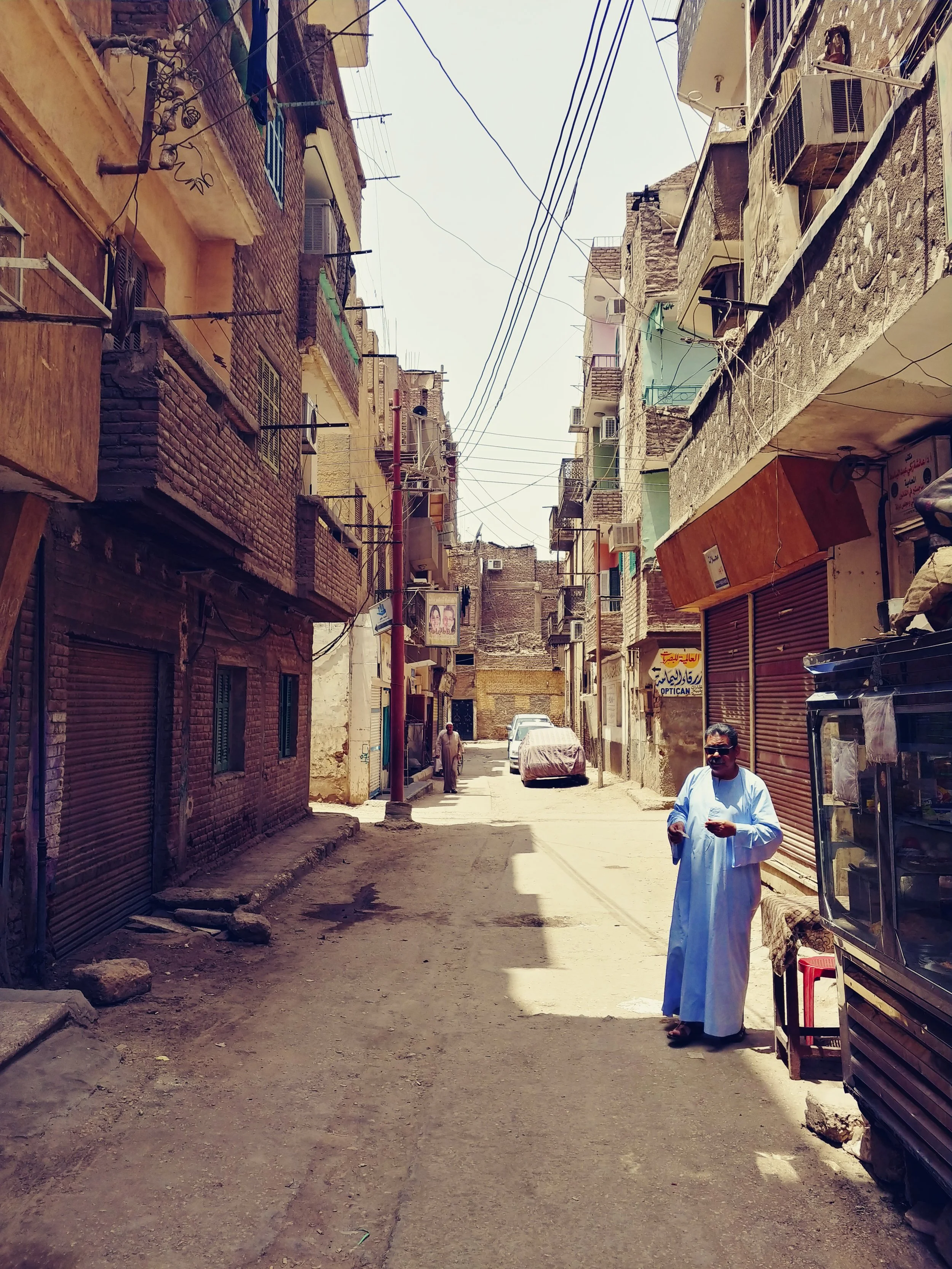 A narrow street in a residential neighborhood with multi-story buildings on either side, some with balconies and air conditioning units. There is a man dressed in traditional white clothing standing near a vending machine or small shop, and another p