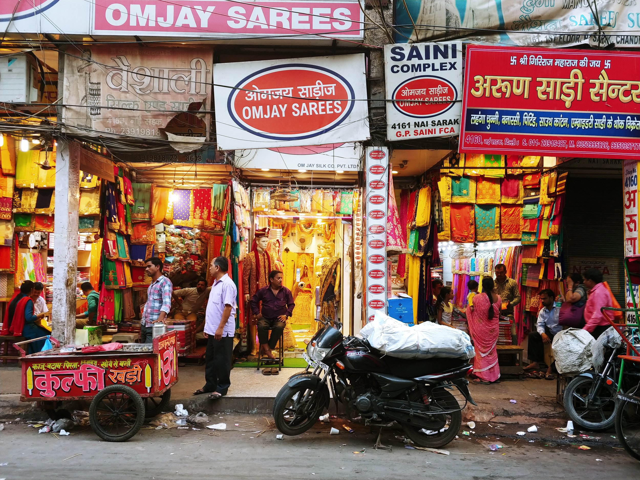 Street market scene with colorful sarees on display, shoppers and vendors, motorcycles parked in front, and various signs in Hindi and English advertising shops named 'Omjay Sarees' and others.