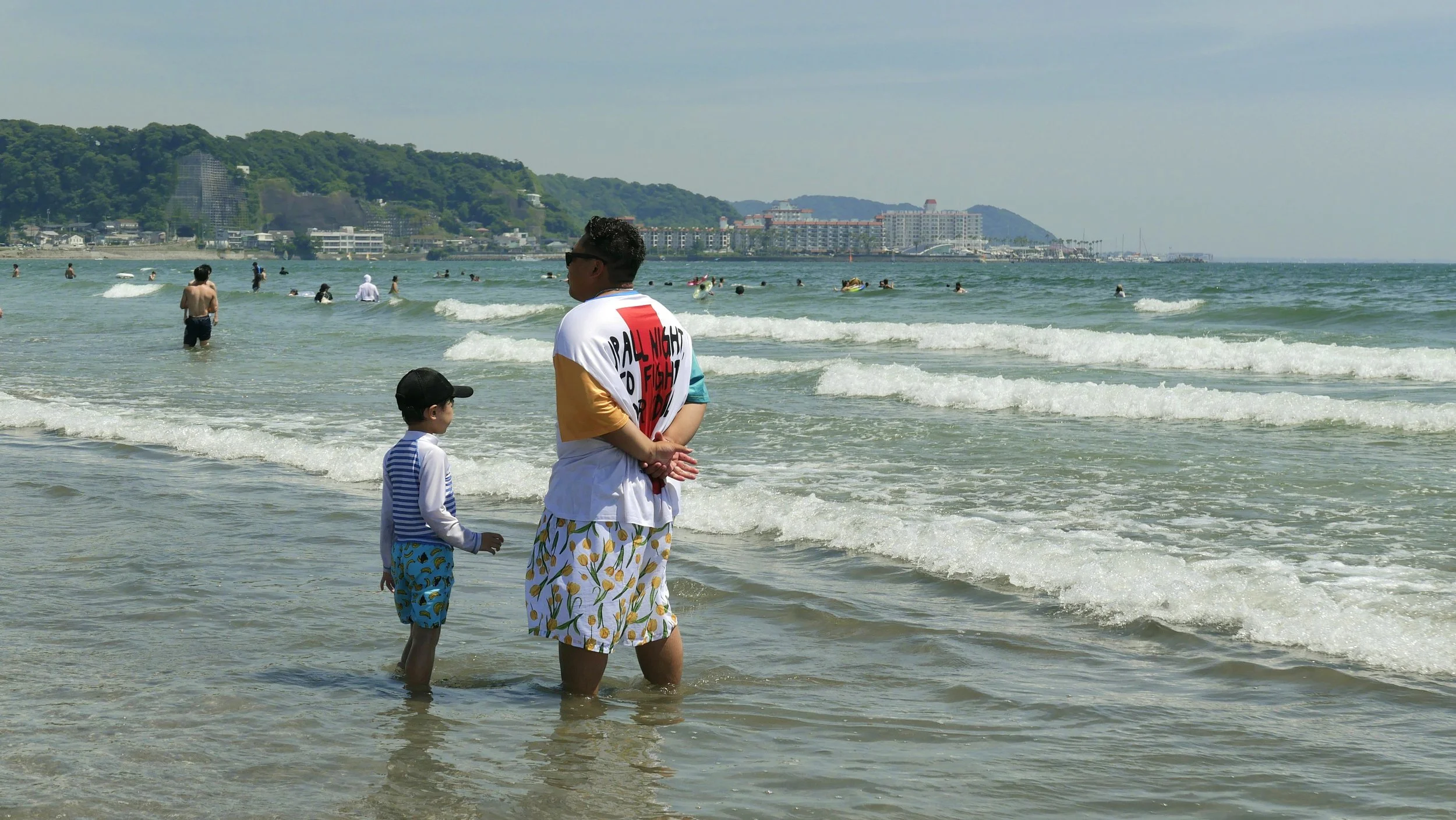 A woman and a young boy standing on the beach near the water, with people swimming and surfing in the background and a cityscape with buildings and green hills beyond.