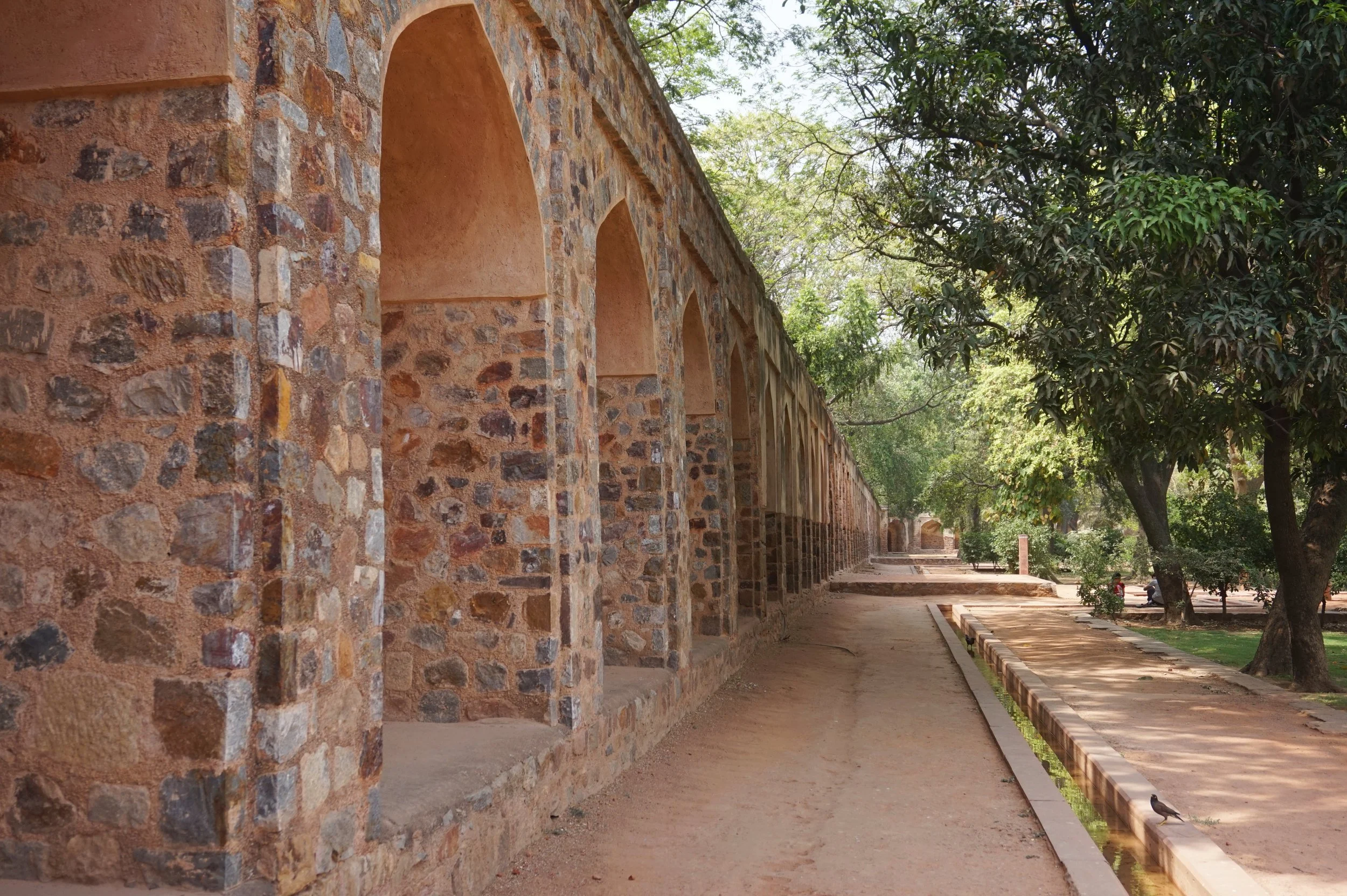 A long stone wall with arches and a pathway beside it, shaded by large trees, in a park-like setting.