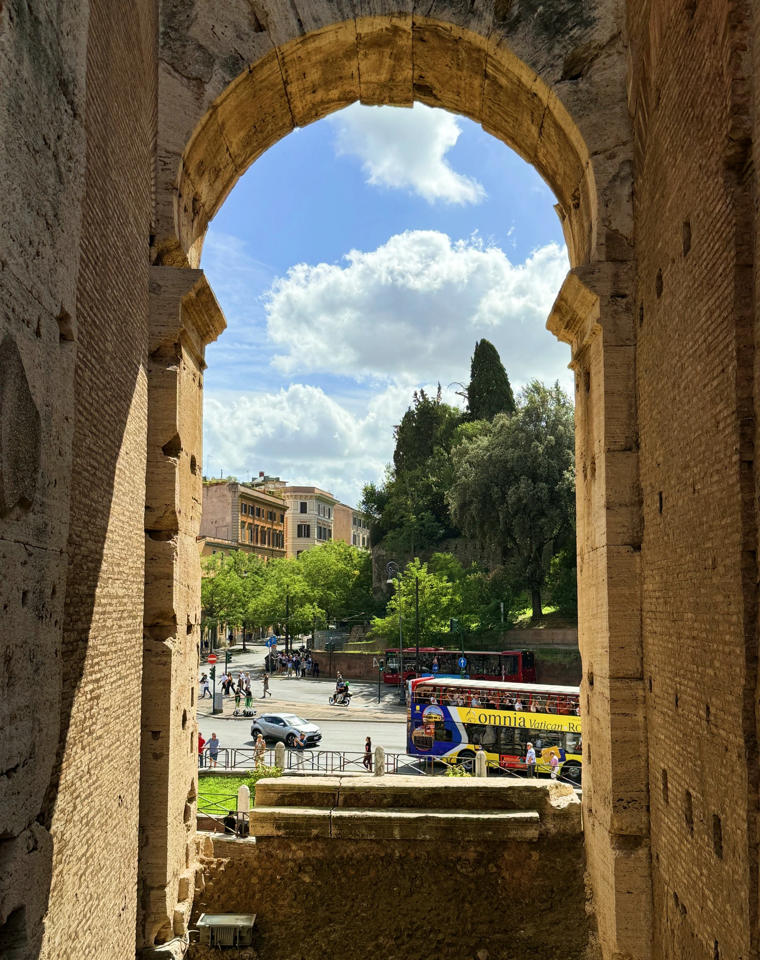 View through an ancient stone archway showing a modern city street with people, cars, and buses, and green trees under a blue sky with clouds.