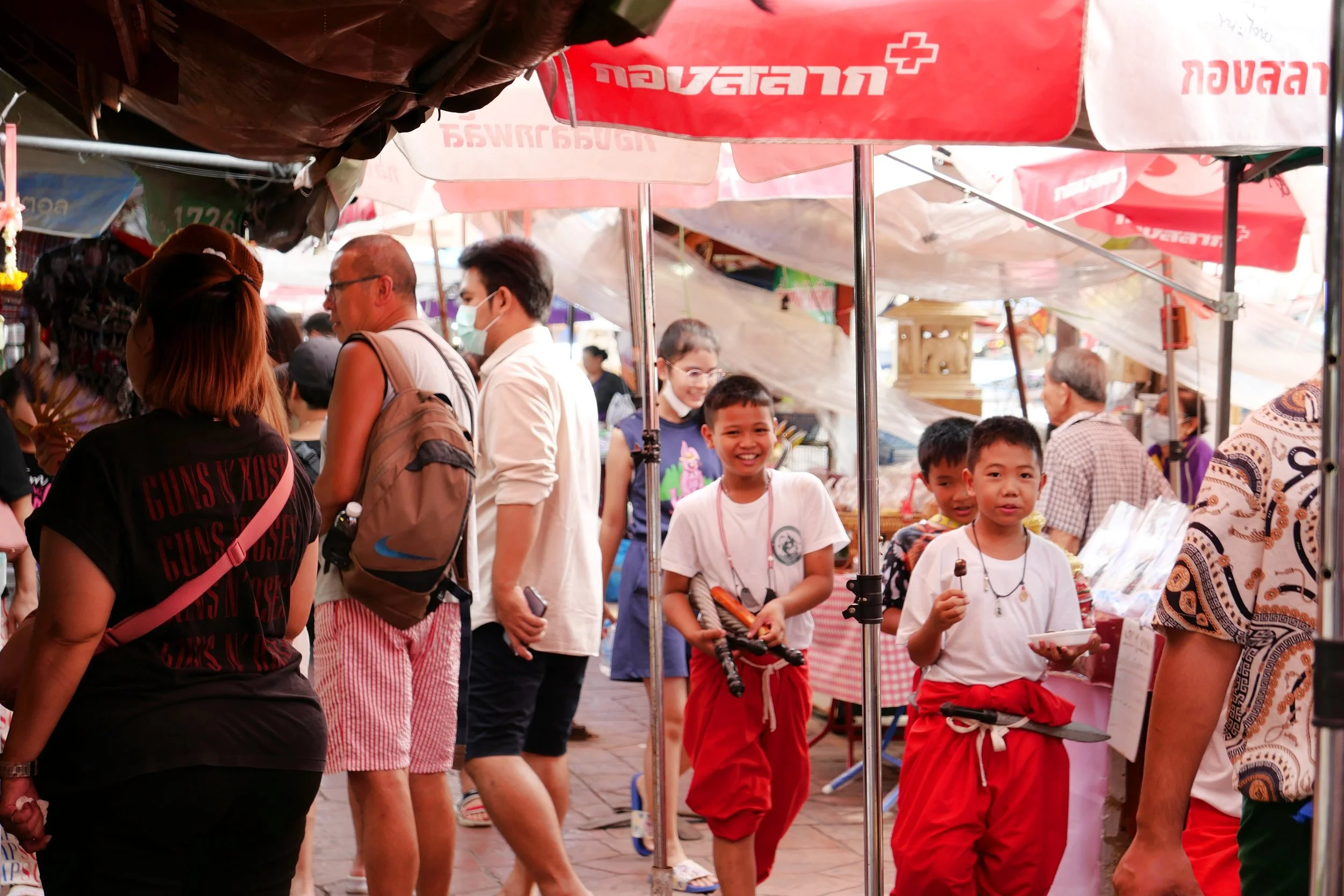 Crowd of people at an outdoor market with vendors and colorful umbrellas, including children smiling and a woman with a brown hat.