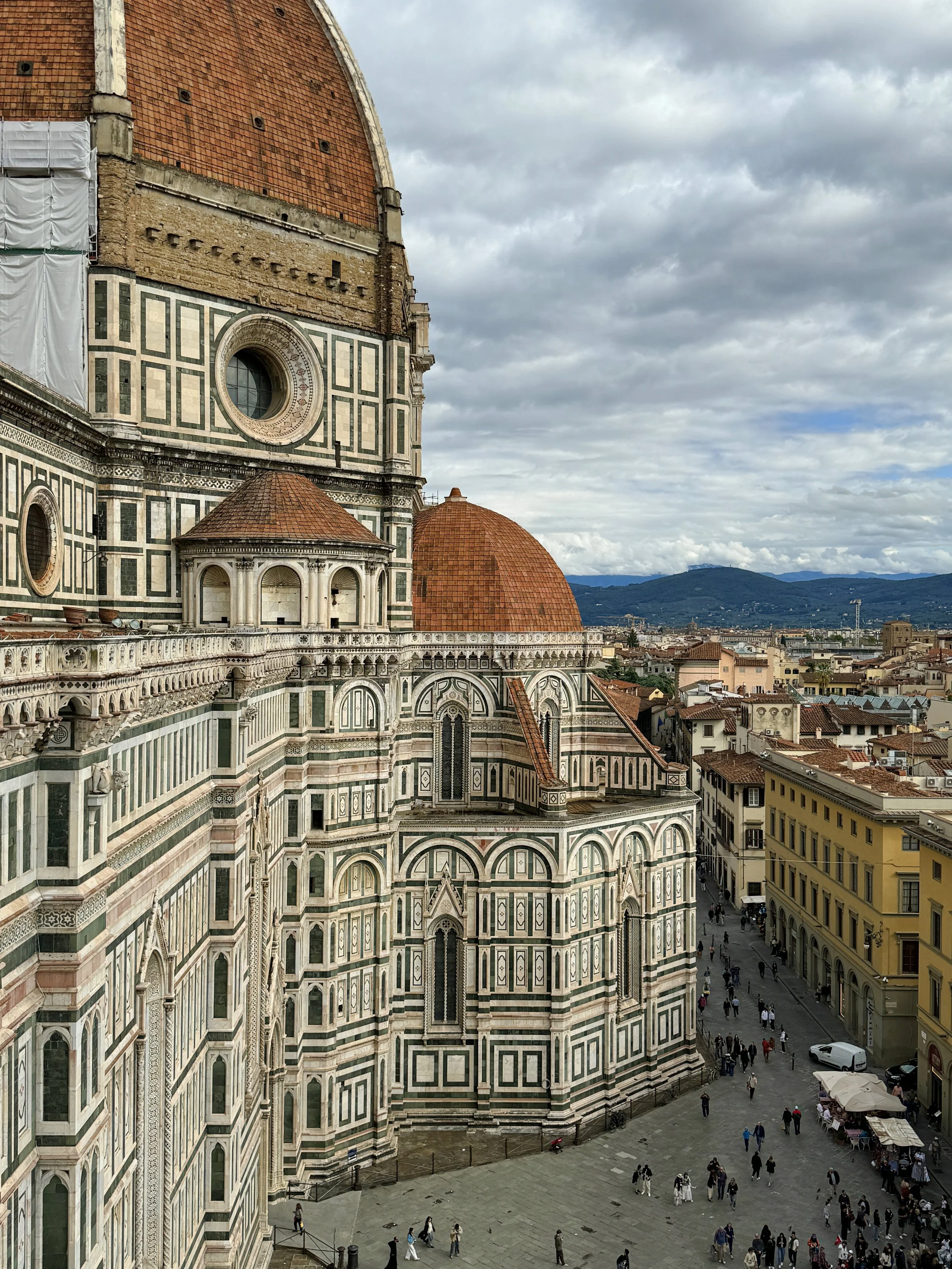 The elaborate facade of the Cathedral of Santa Maria del Fiore in Florence, Italy, with intricate green, white, and pink marble patterns, arched windows, and stone details, surrounding a bustling city square filled with people and street cafes under 