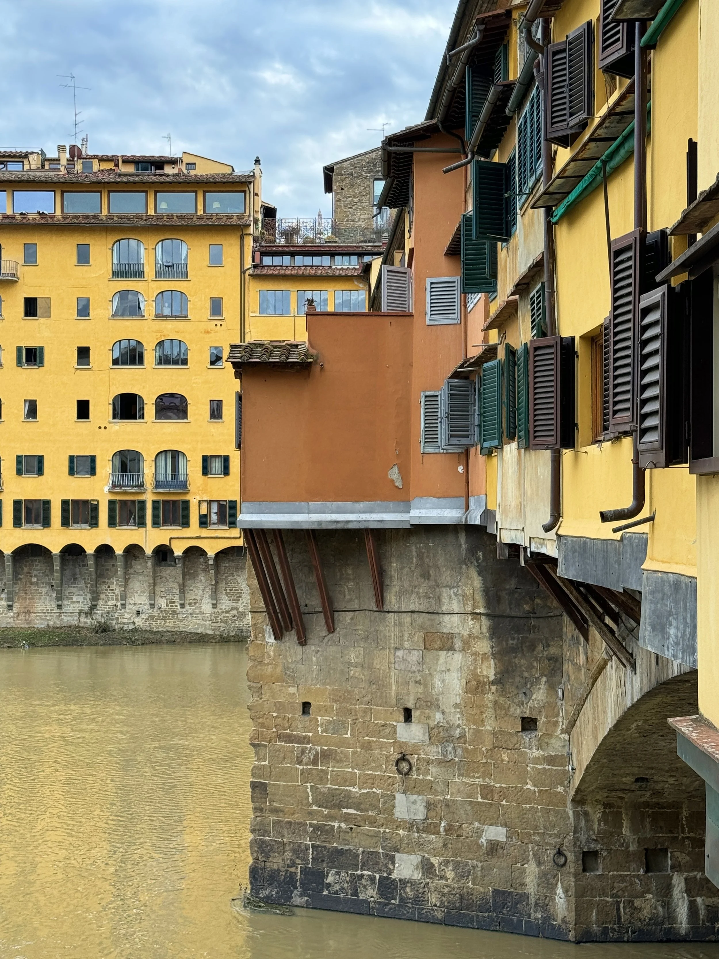View of colorful buildings along the riverbank, with windows, shutters, and a stone bridge supporting the structures, situated next to a body of water under a partly cloudy sky.