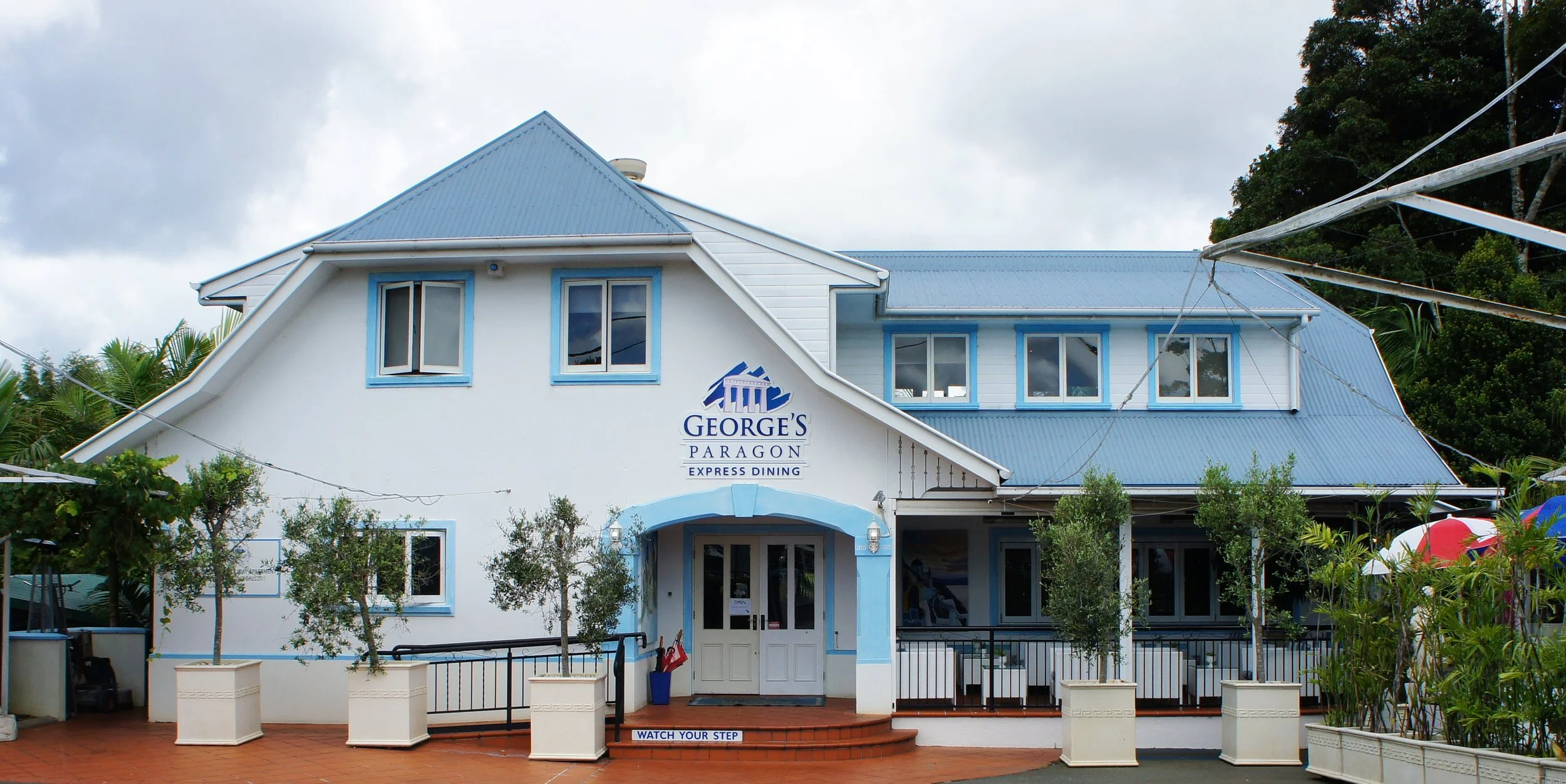 A two-story building with a blue metal roof and white exterior walls, labeled George's Paragon Express Dining, surrounded by trees and plants, with a paved area in front and a sign that says 'Watch Your Step' at the entrance.