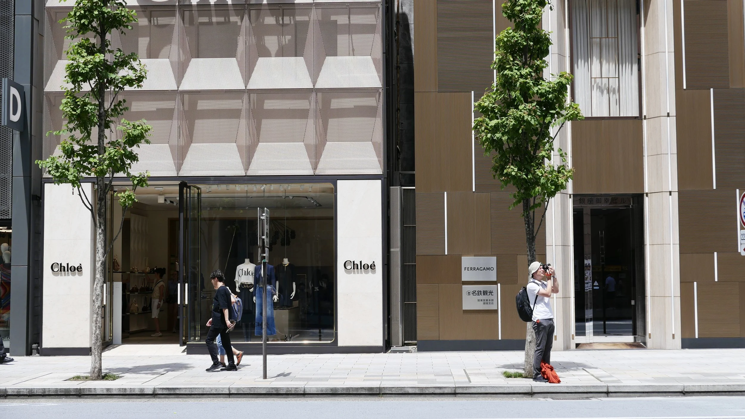 City street scene with two trees, a store with a Chloé sign and other brand signs, people walking, and a woman taking a photograph.