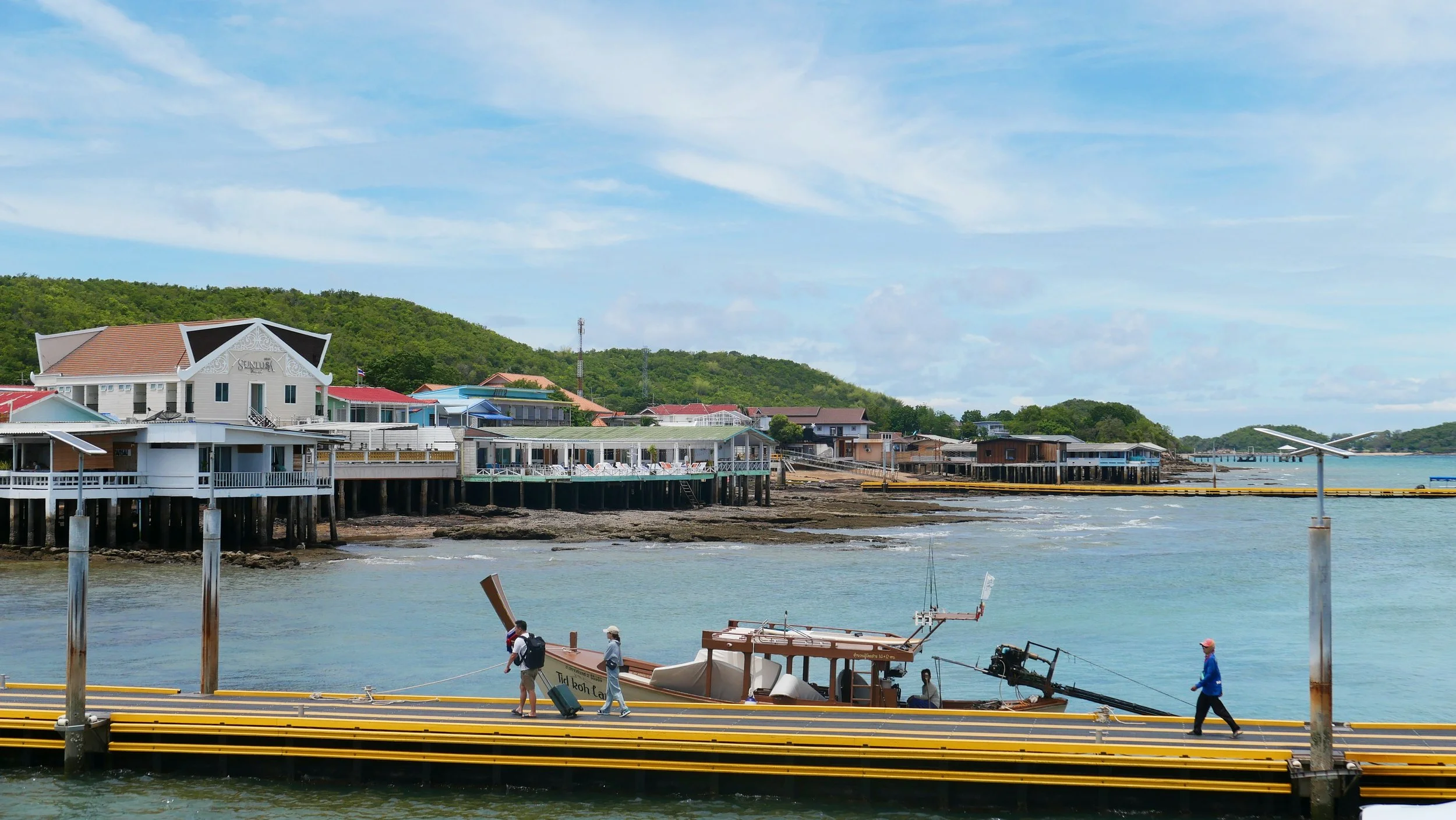 A coastal scene with colorful buildings on stilts along the shoreline, boats on the water, and a bridge in the distance under a partly cloudy sky.