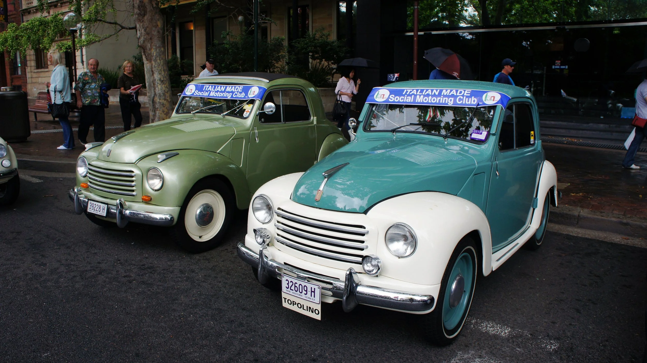 Two vintage cars on display, both with signs indicating they are part of the Italian Made Social Motoring Club, parked on a city street with people walking and standing nearby, some holding umbrellas.