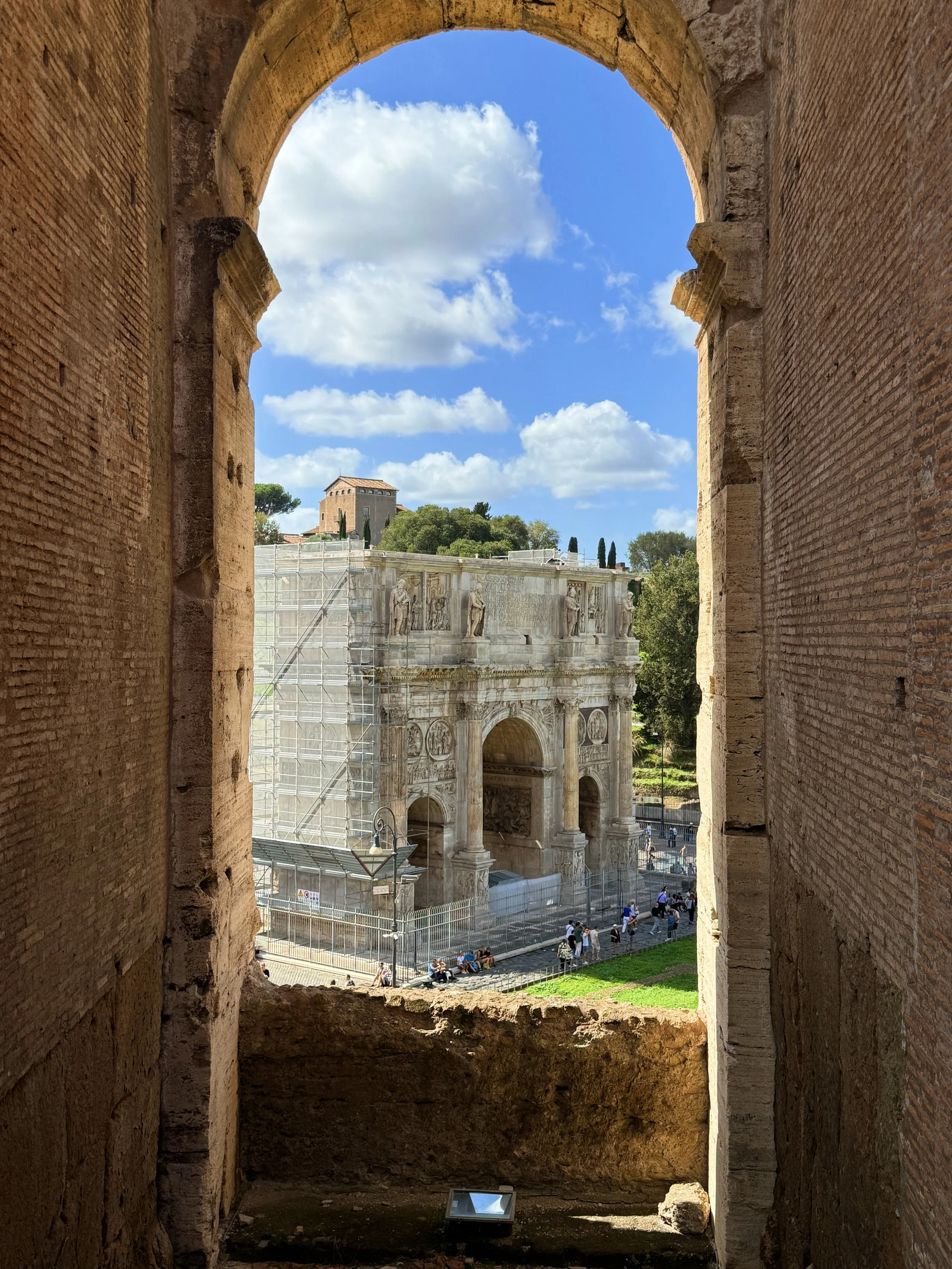 View of an ancient Roman triumphal arch seen through a brick window, with a blue sky, white clouds, and a historic building with statues and trees in the background.