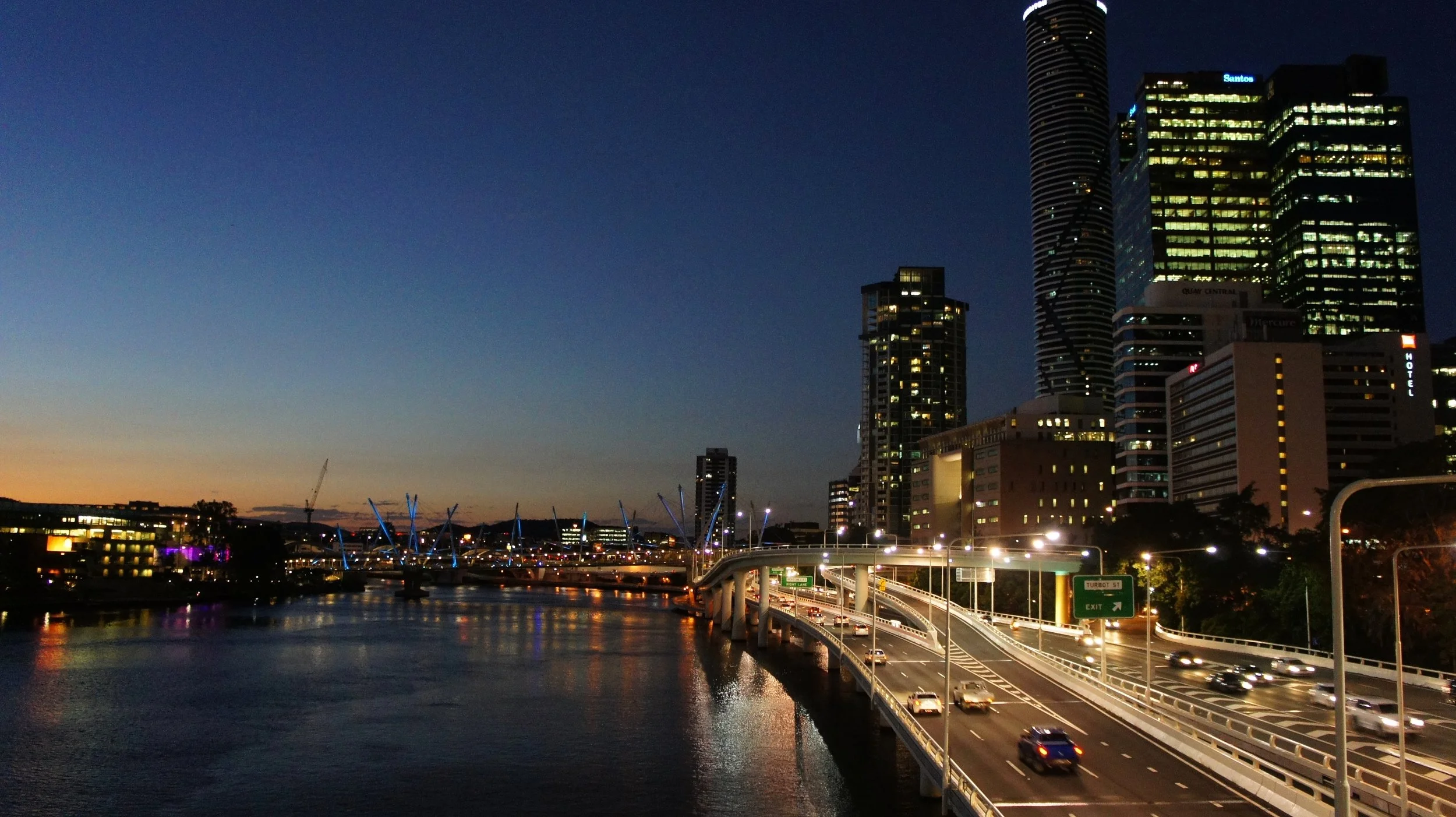 City skyline at dusk with illuminated skyscrapers, a bridge with traffic, and buildings reflected in a river.