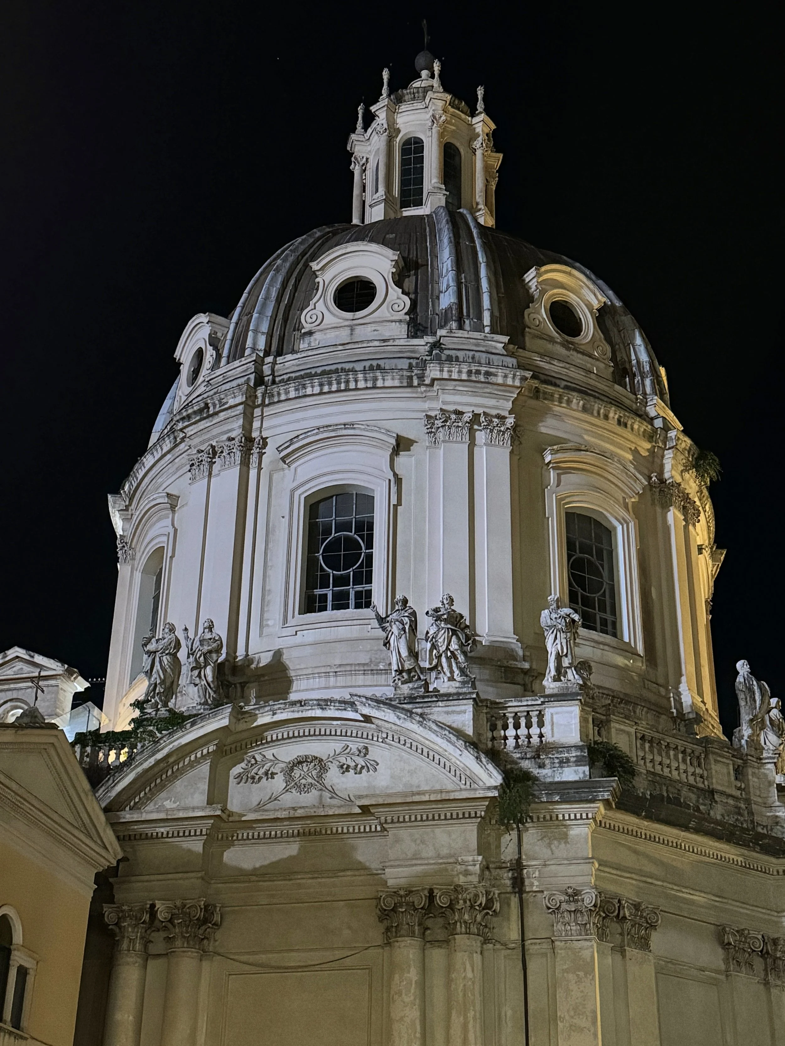 Night-time view of an ornate baroque-style building, possibly a church, with detailed sculptures and large arched windows, illuminated against a dark sky.
