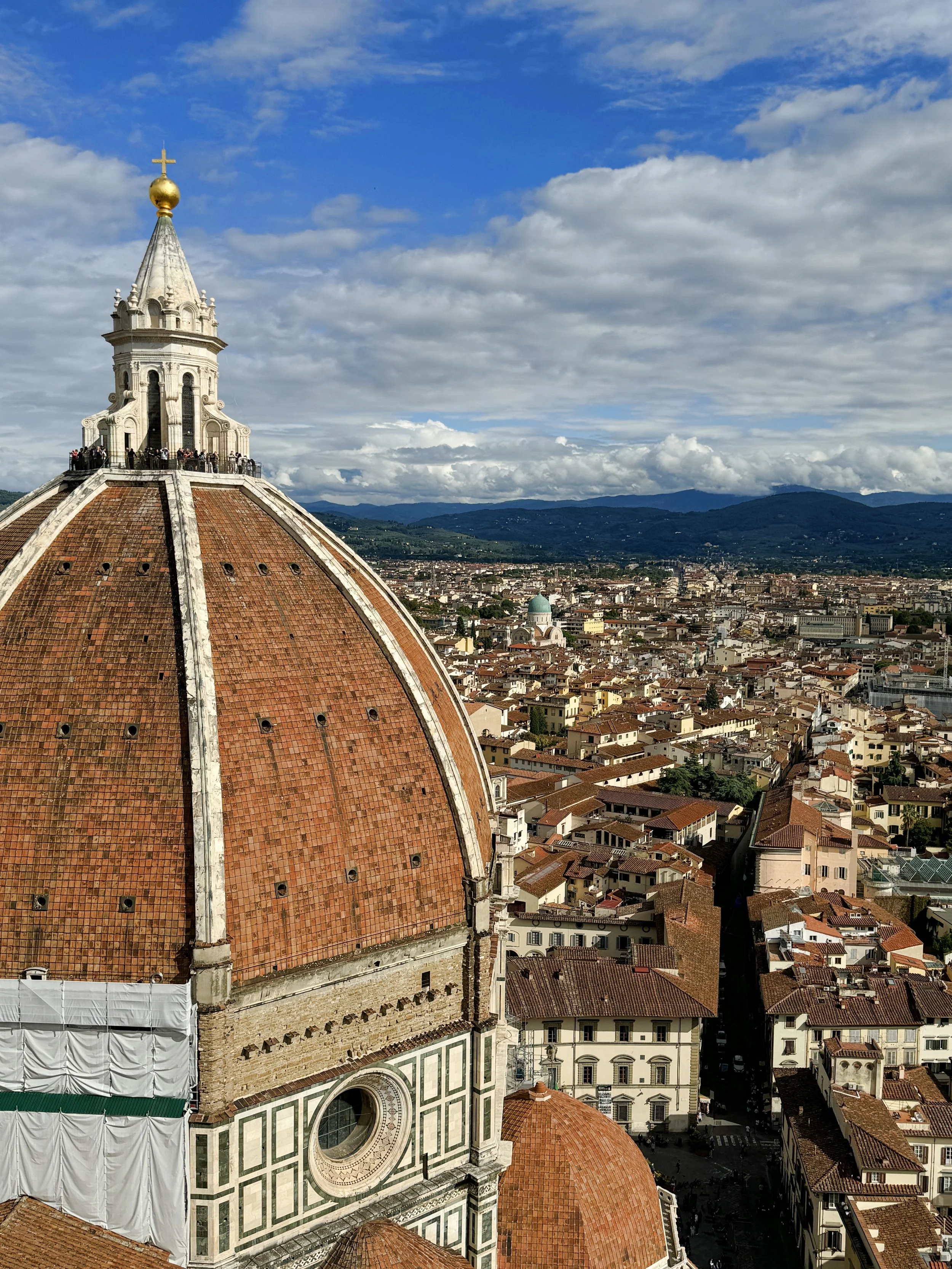 Aerial view of Florence, Italy, featuring the dome of Florence Cathedral with a gold cross and orb on top, city rooftops, and distant mountains under a partly cloudy sky.