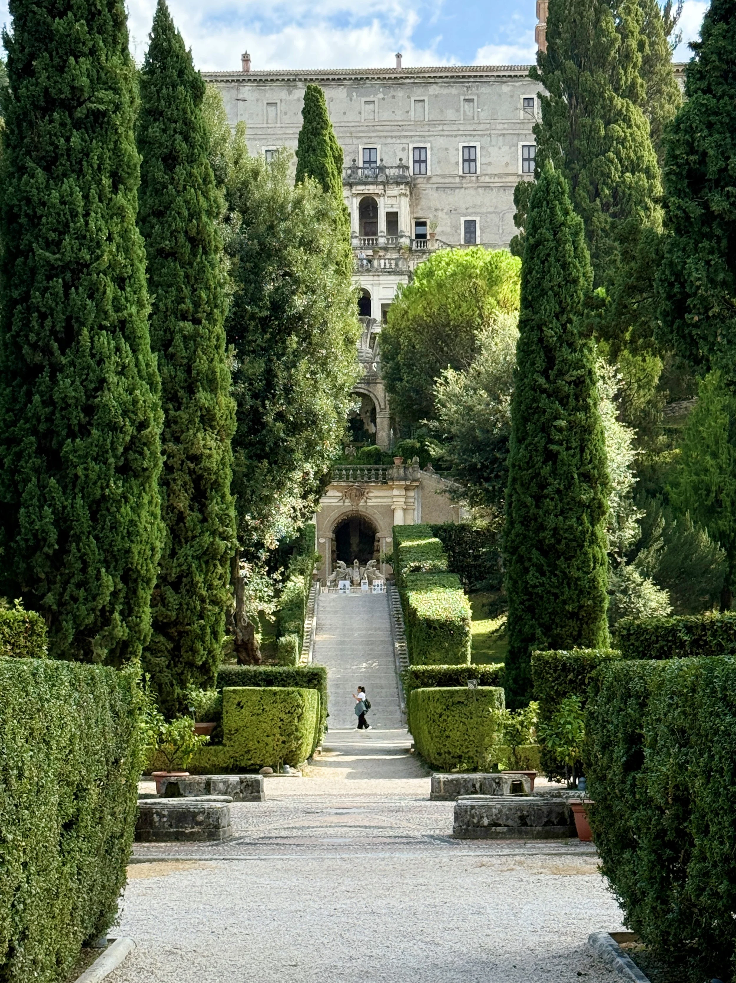 A historic European castle or villa surrounded by a lush green garden with tall cypress trees, trimmed hedges, and a stone staircase leading up to the building.