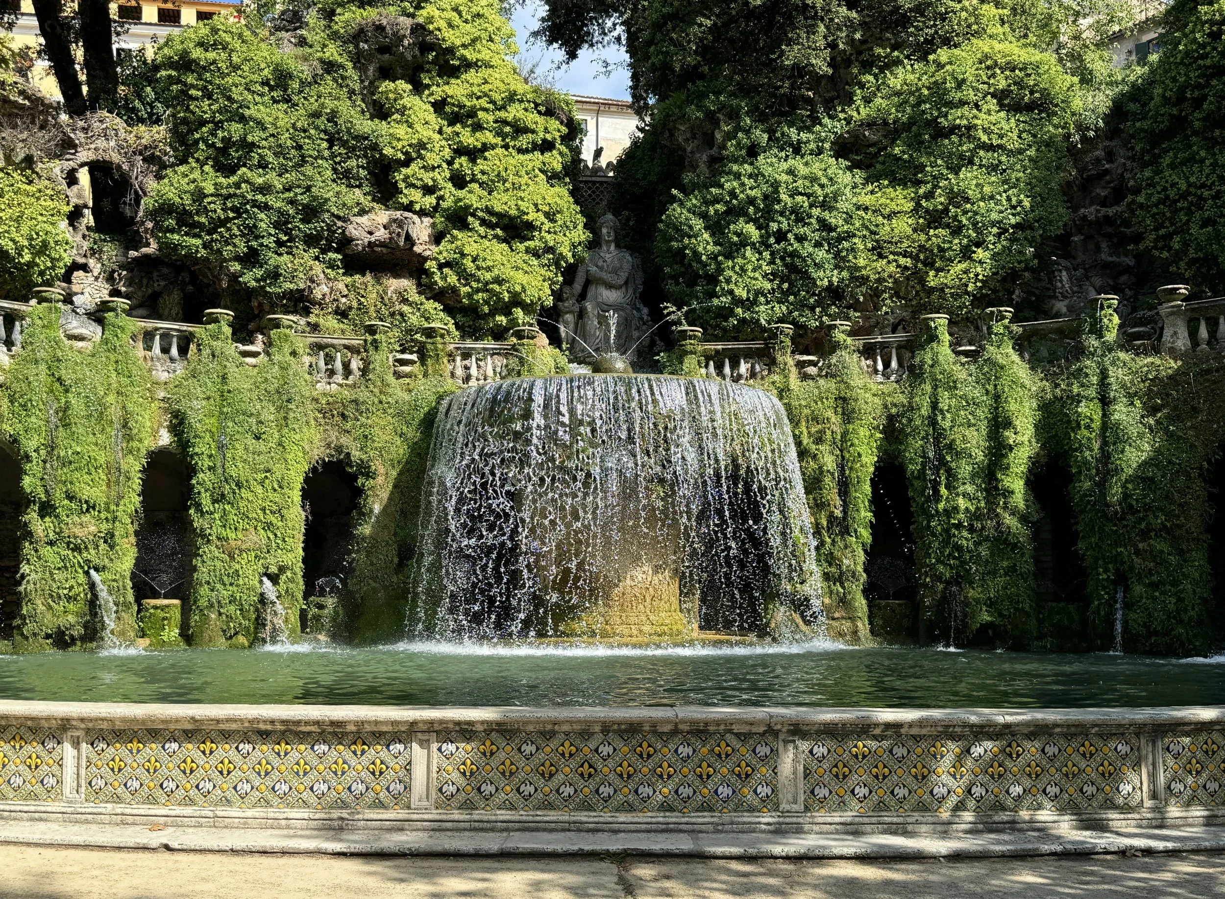 A fountain with a sculpture of a woman at the center, surrounded by lush green trees, with water cascading from the sculpture into a pool below. There is a decorative tiled wall at the base of the fountain.