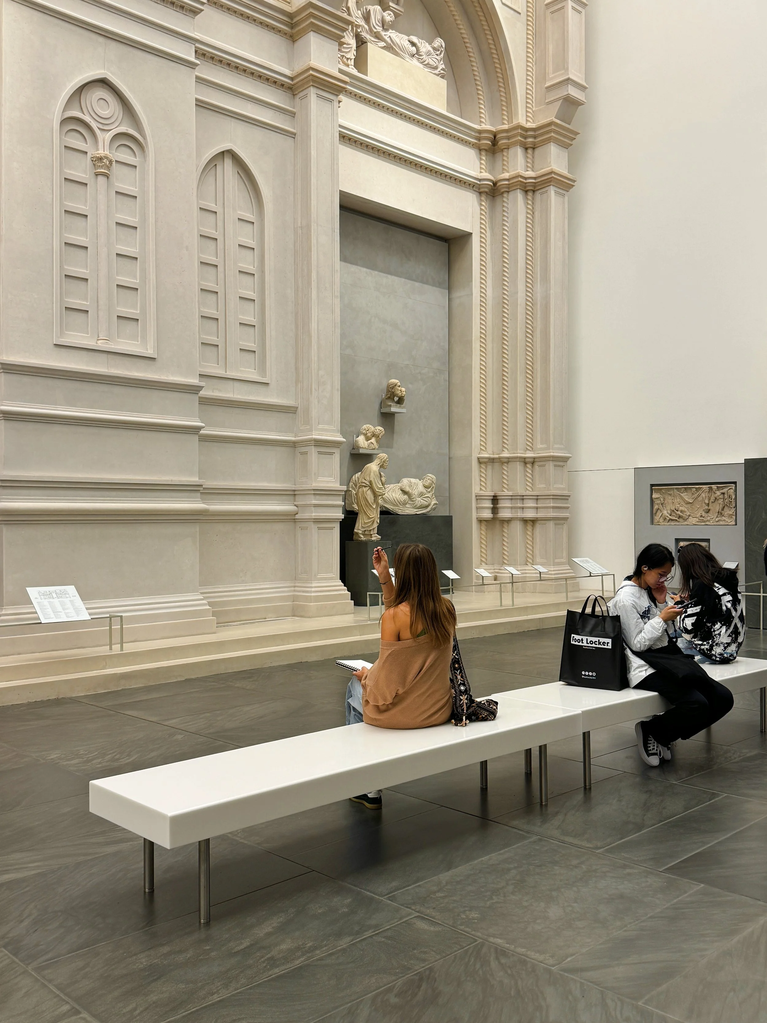 People sitting on a white bench in a museum with classical sculptures and intricate architectural details on the wall.