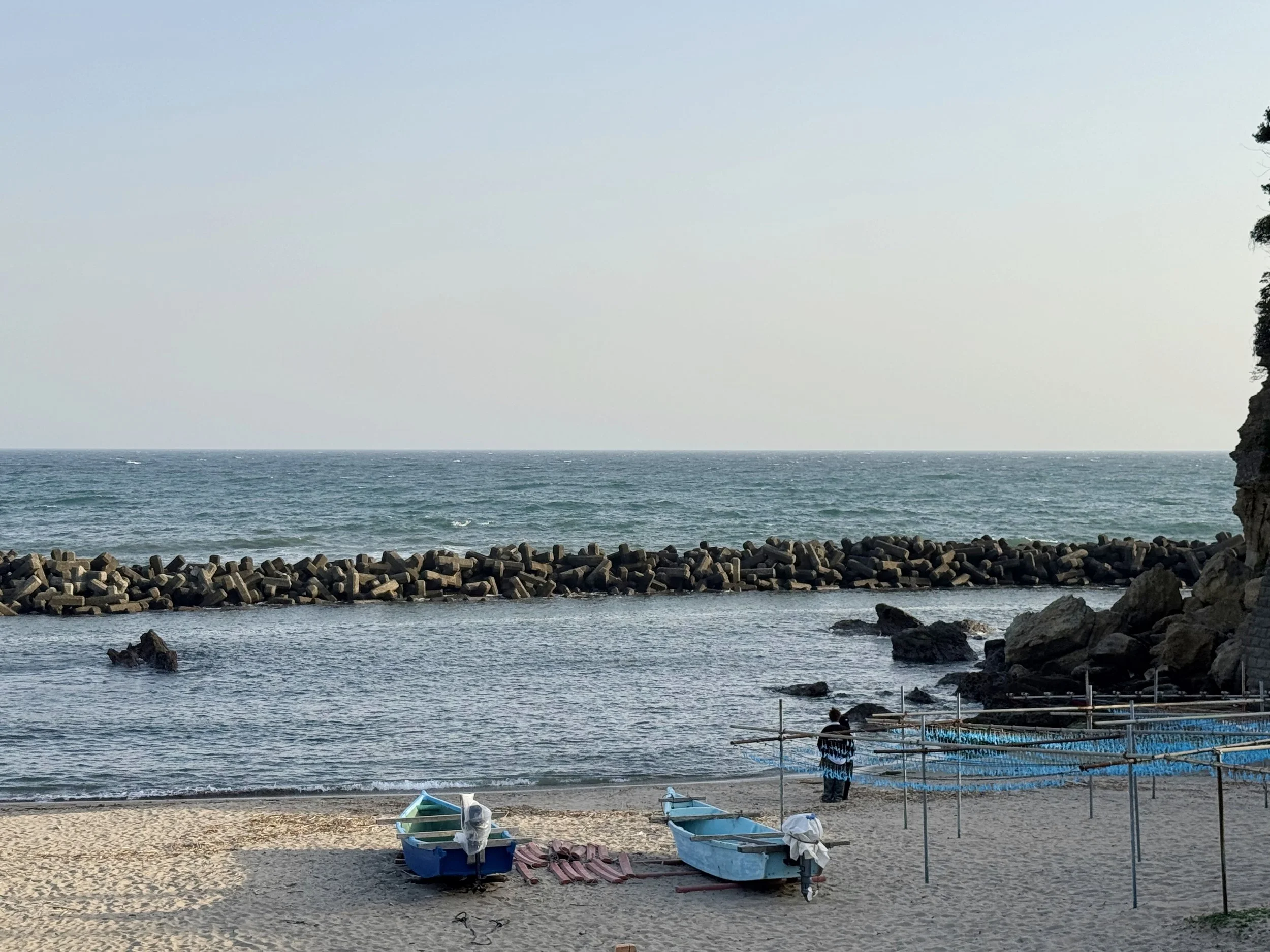 View of a beach with small boats on the sand, a shoreline with rocks, a breakwater extending into the ocean, and a foggy sky over the water.