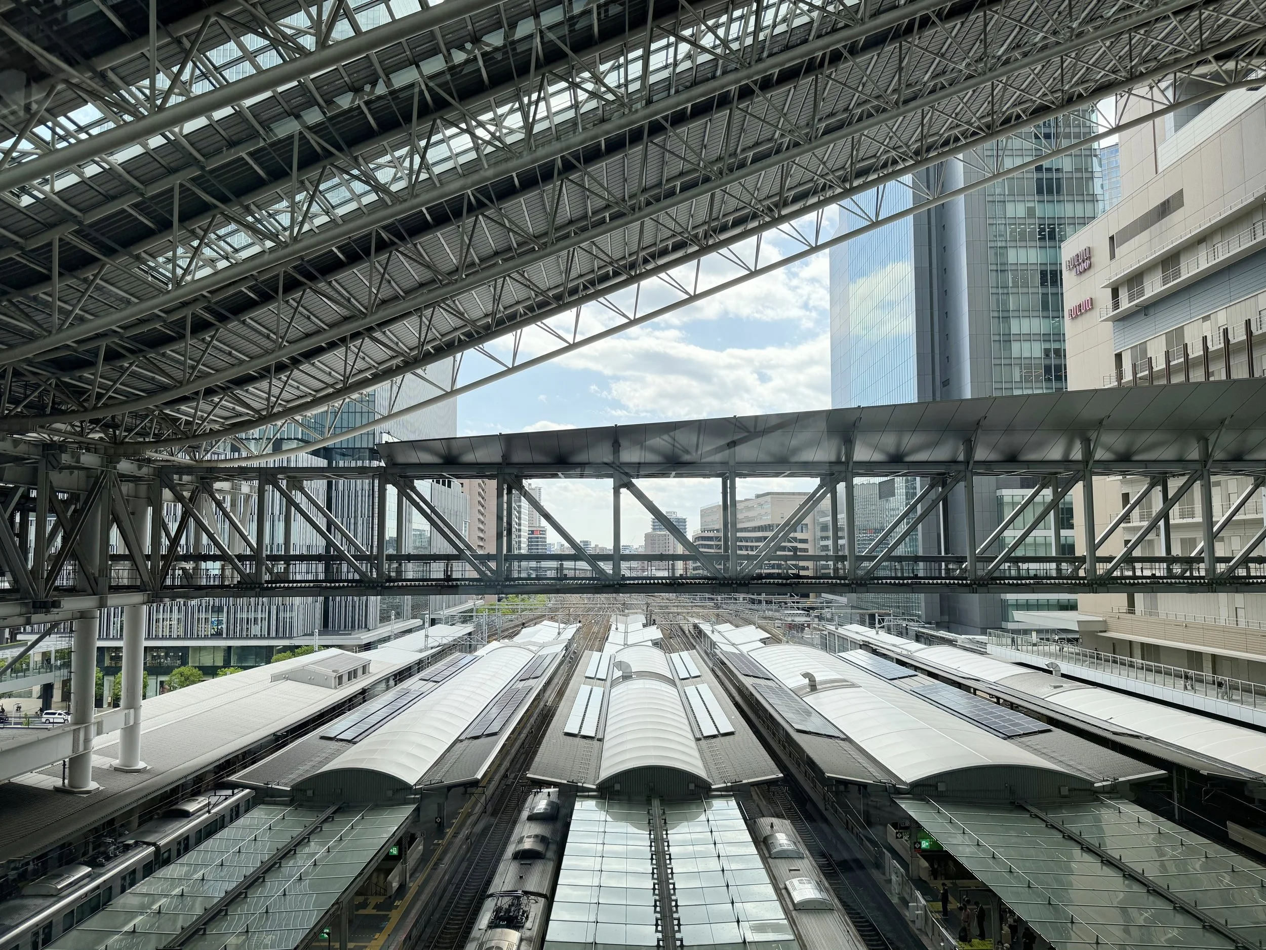 View of an indoor train station with multiple train tracks, platforms, and a modern architectural ceiling structure with metal beams and glass. Tall buildings are visible outside through the glass windows.