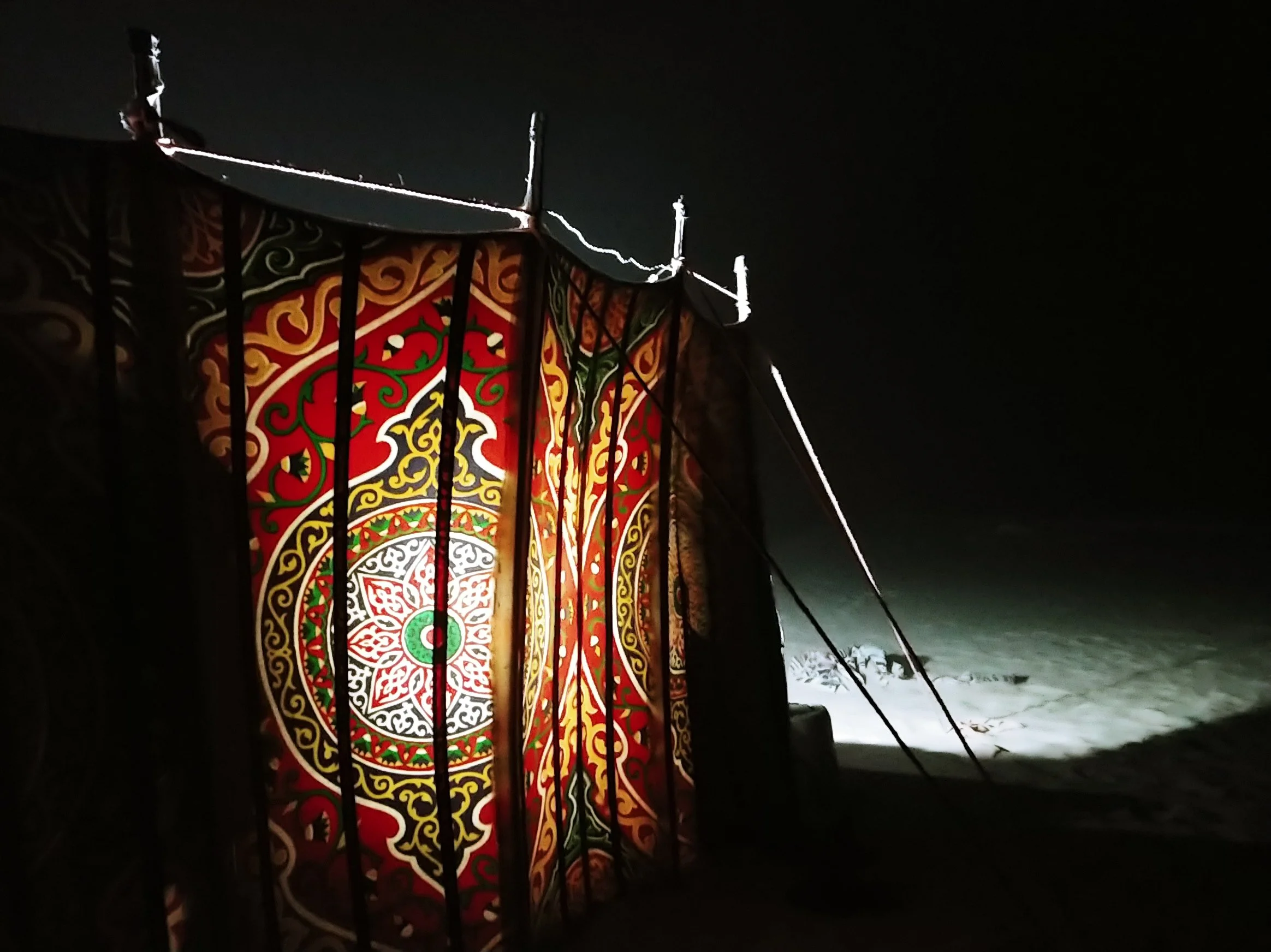 Colorful lantern with intricate patterns glowing at night, secured behind metal bars, with snow on the ground.