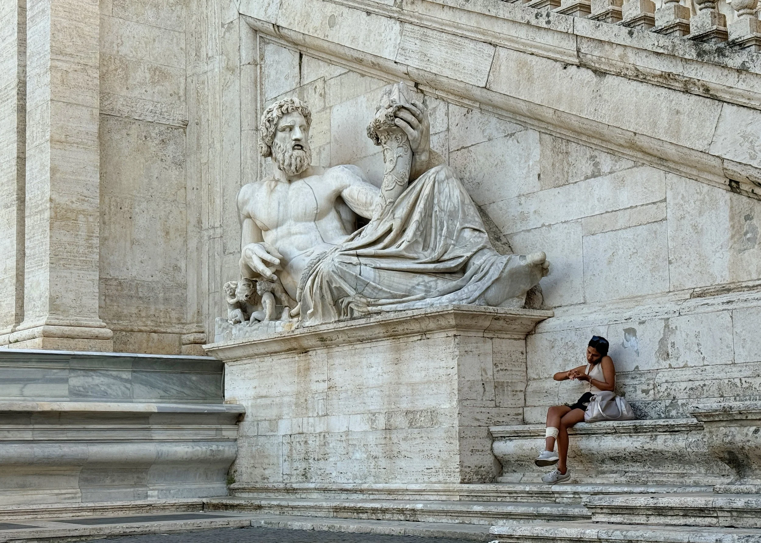 A white marble statue of a reclining man with a beard, holding a helmet in his left hand. The statue is mounted on a stone wall with classical architectural elements. Two women are sitting below the statue, one with crossed legs and engrossed in her 