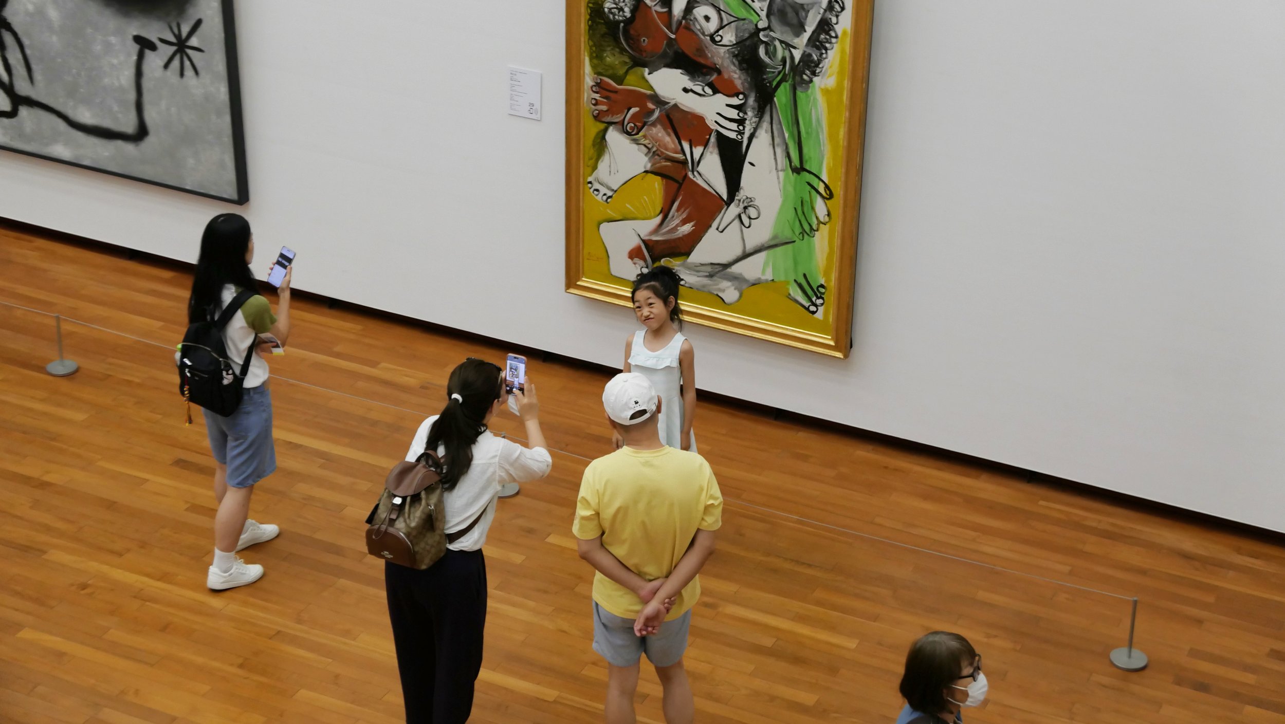 A group of people, including children, are observing and taking photographs of a colorful abstract painting in an art gallery.
