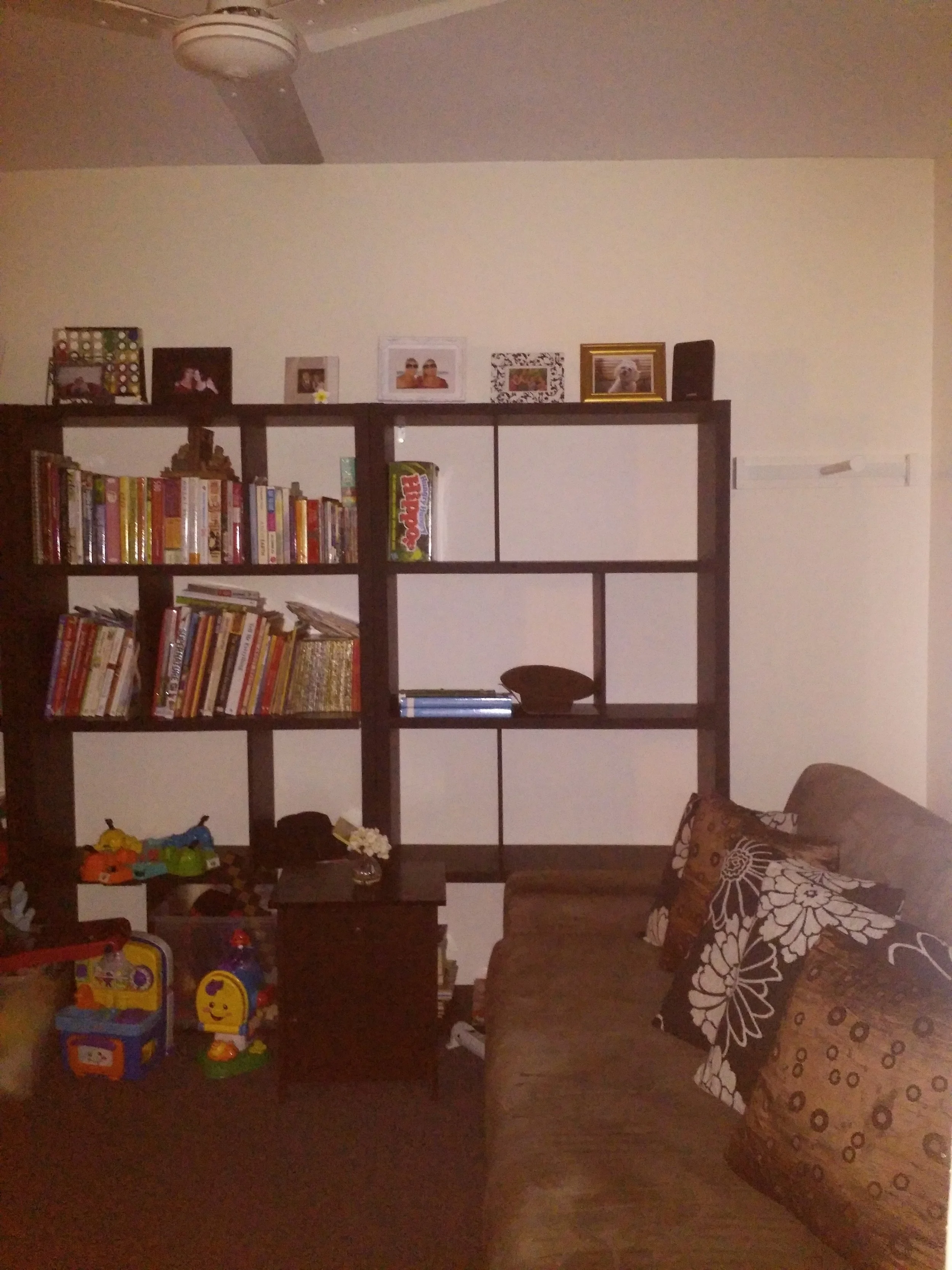 Living room with a bookshelf filled with books, framed photos on the wall, a brown sofa with a patterned pillow, a side table with a small flower vase, and various children's toys on the floor.