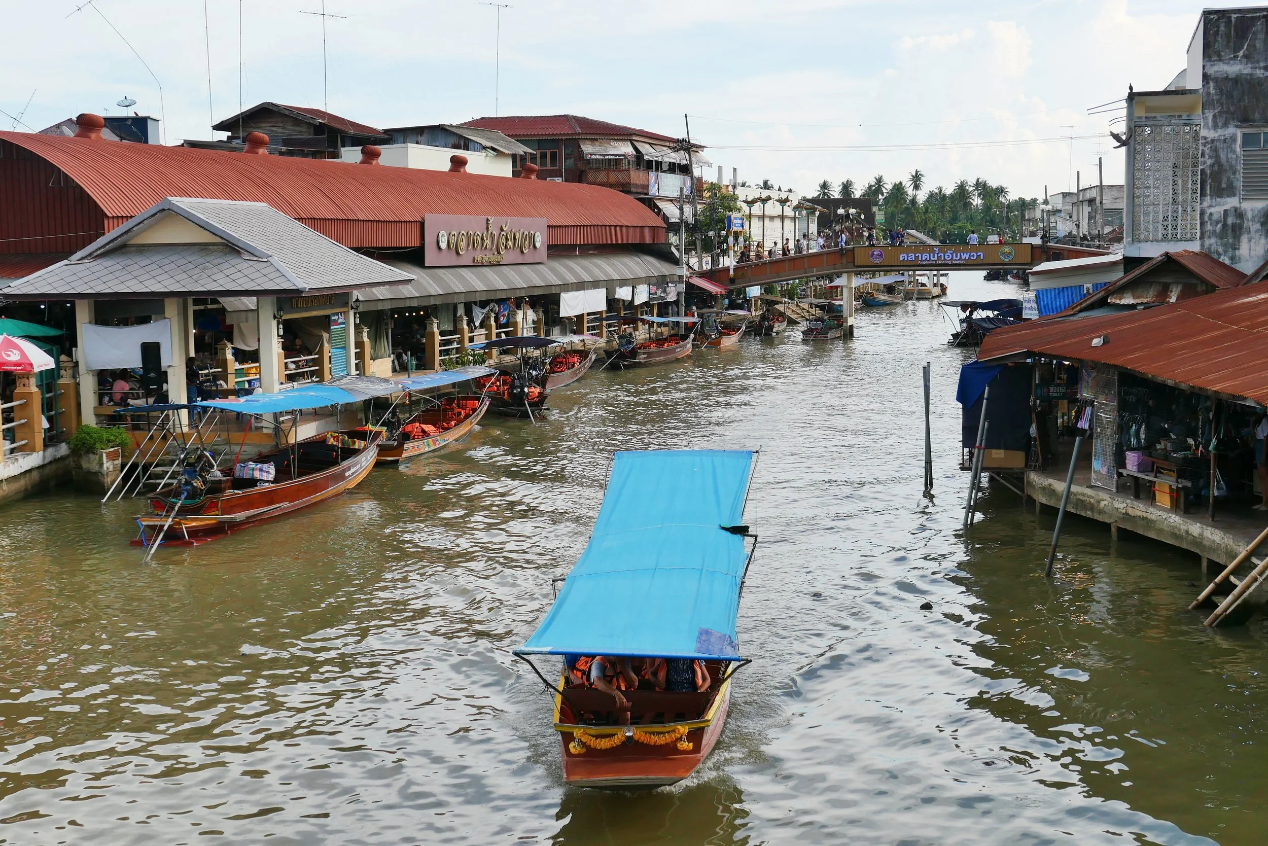A floating market scene with boats on a river, storefronts with signs in a foreign script, and a pedestrian bridge with people, in a tropical setting with palm trees.