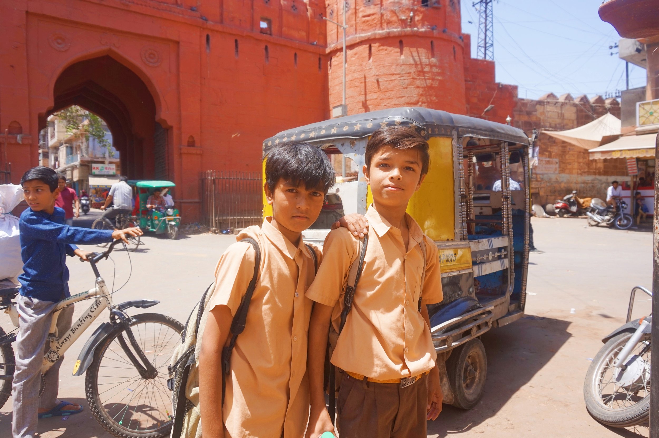Three young boys in school uniforms standing on a busy street in front of a decorated auto rickshaw with a red sandstone fort or palace in the background. One boy on the left rides a bicycle, while two boys on the right pose for the photograph.