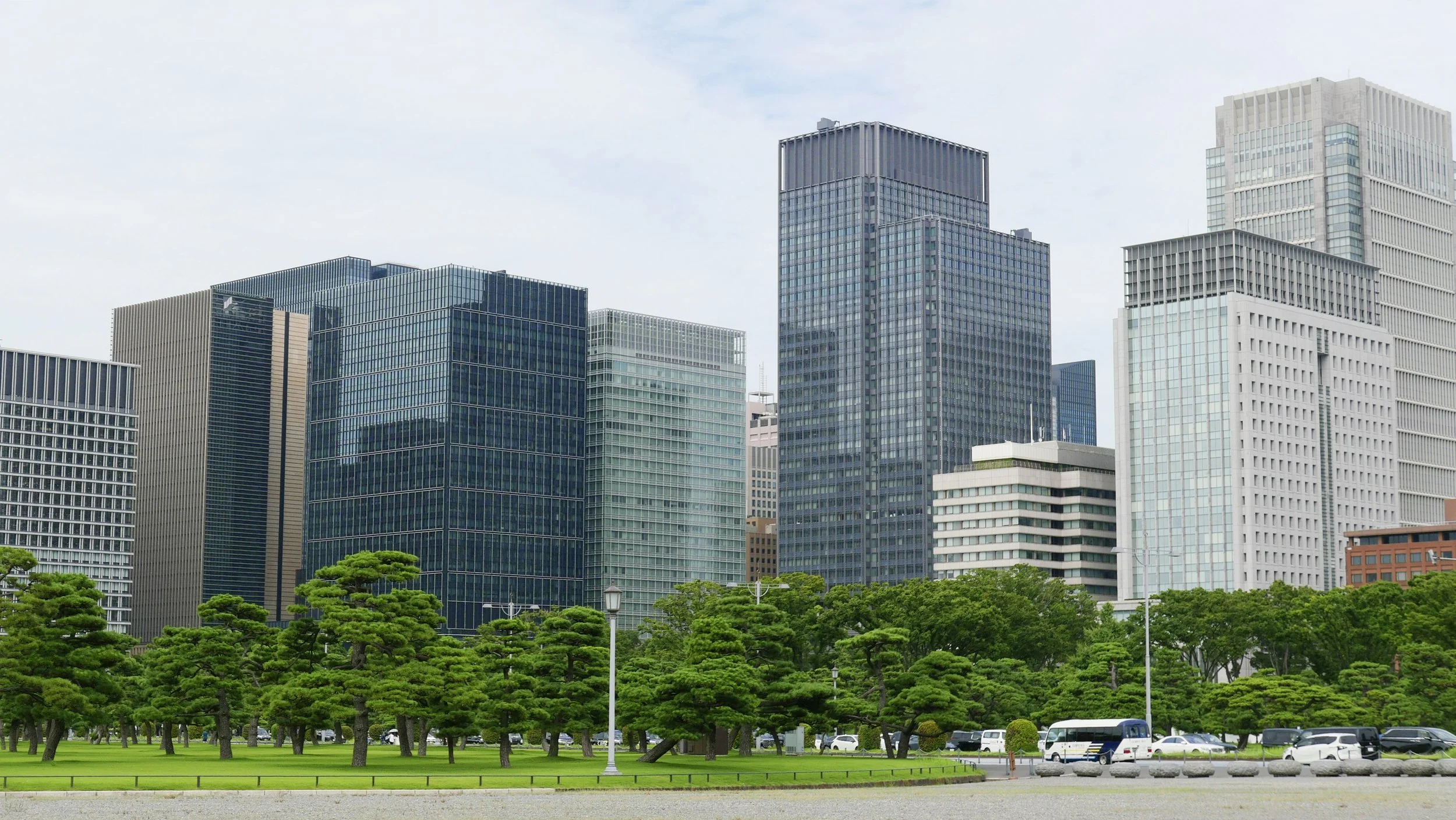 A cityscape with tall modern glass buildings, green trees, and a parking lot in front.
