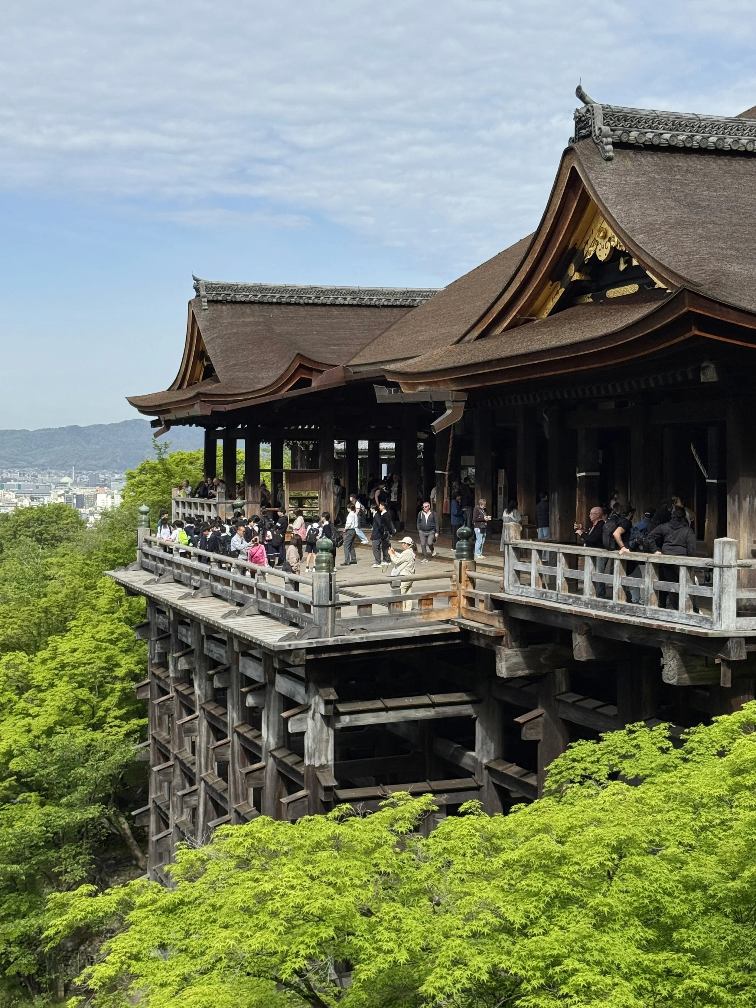 Traditional Japanese temple with wooden structure and ornate roof, surrounded by lush green trees, overlooking a distant cityscape under a cloudy sky.