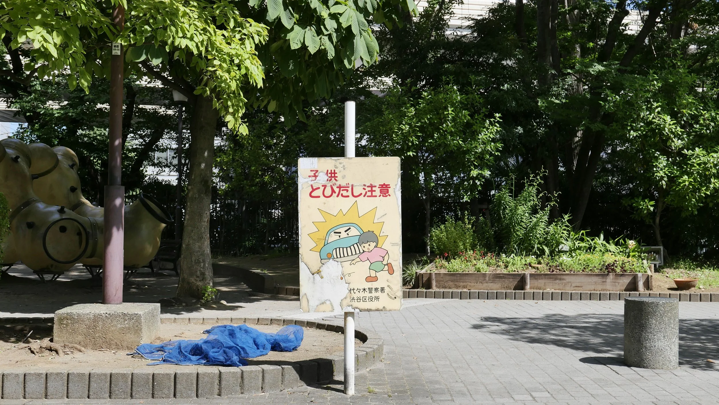 A park with green trees, a sign with cartoon characters warning about falling objects, and playground equipment including large animal-shaped structures in the background.