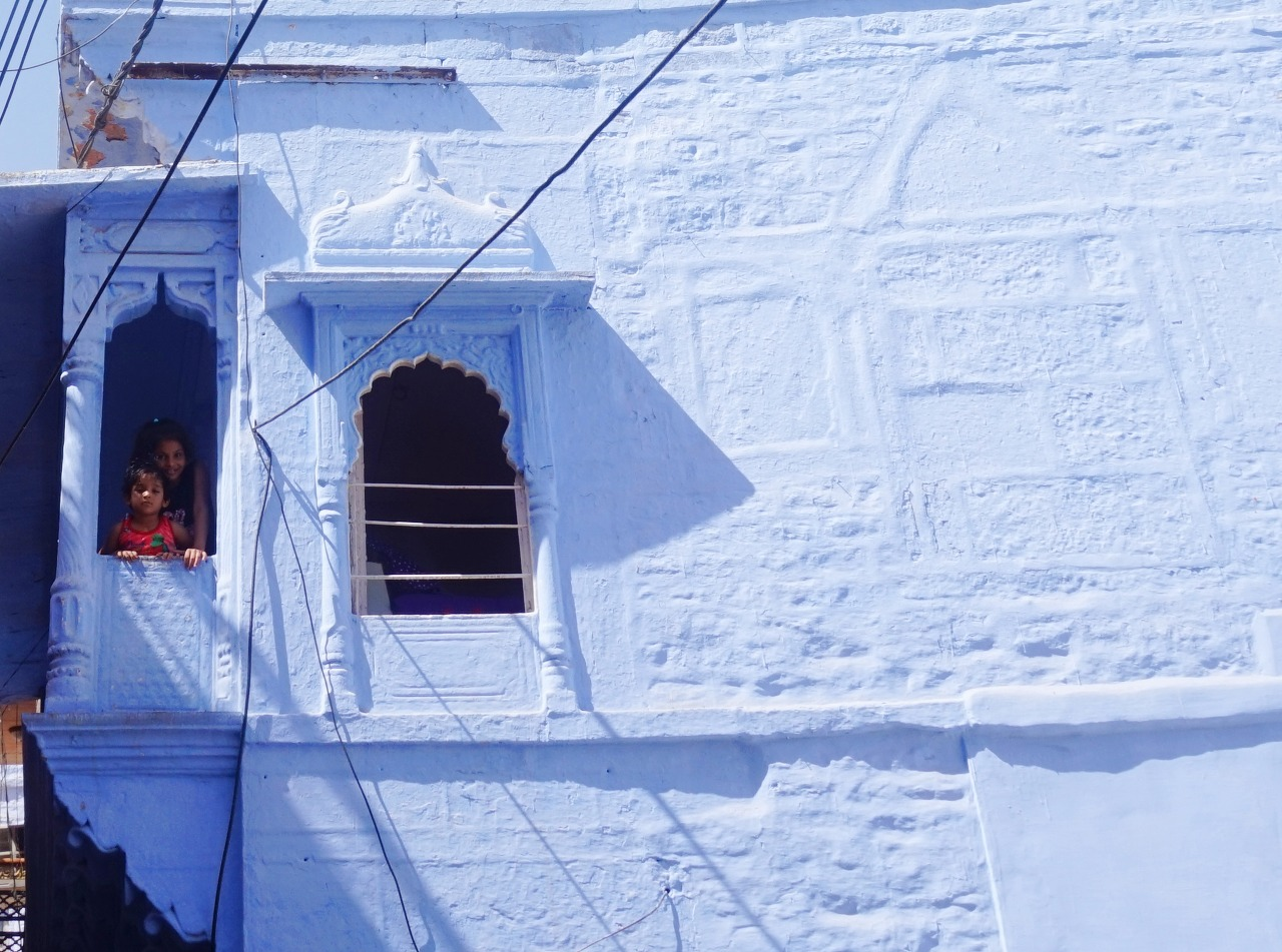 Two children, a young girl and an older girl, standing on a small balcony of a white building with ornate window frames and decorative architectural elements in a coastal town.