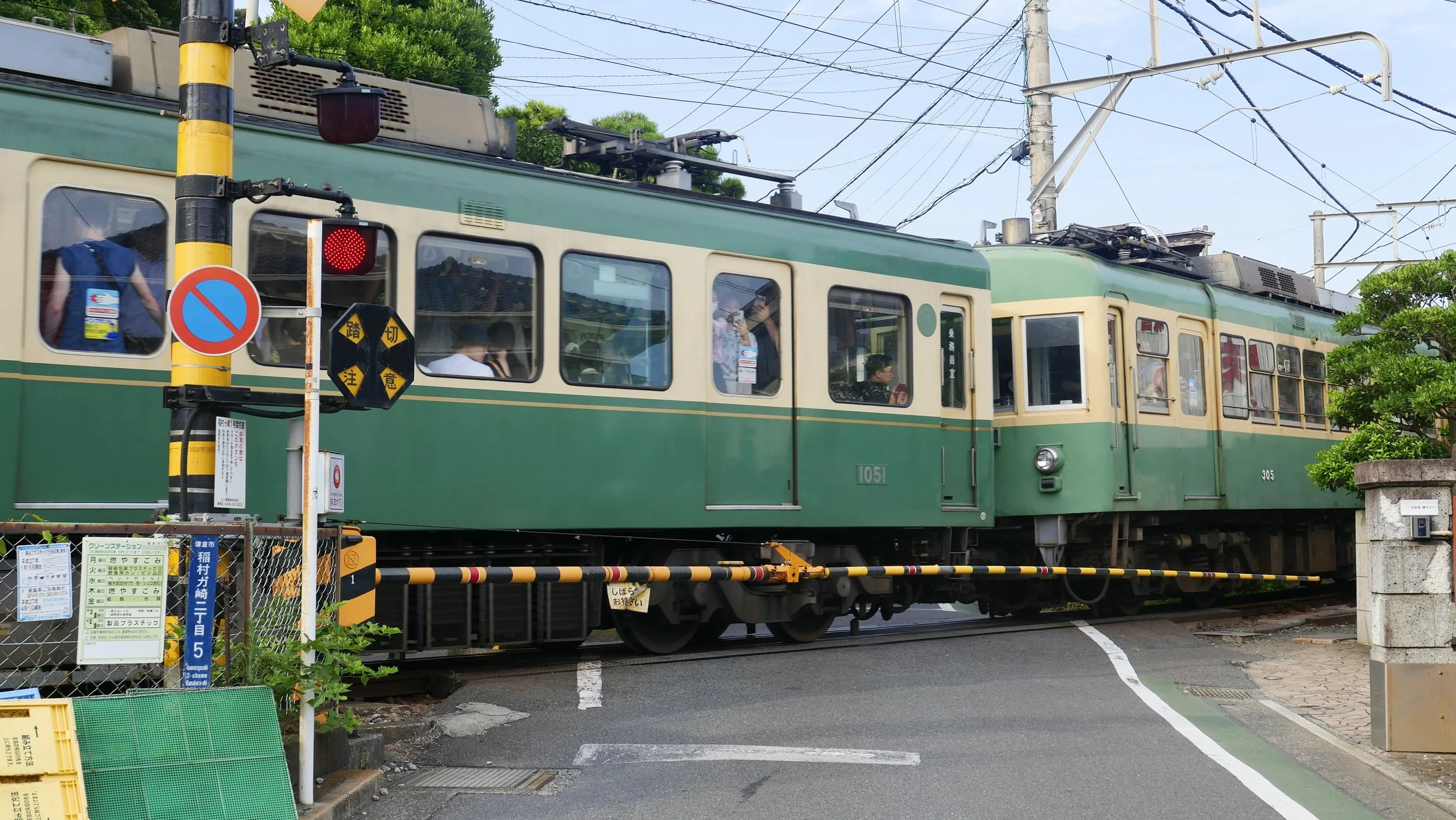 A vintage green and cream-colored train on tracks, with overhead electrical wires and a signal light showing red. There are various signs and forms in Japanese in the foreground, with some trees and a blue sky in the background.