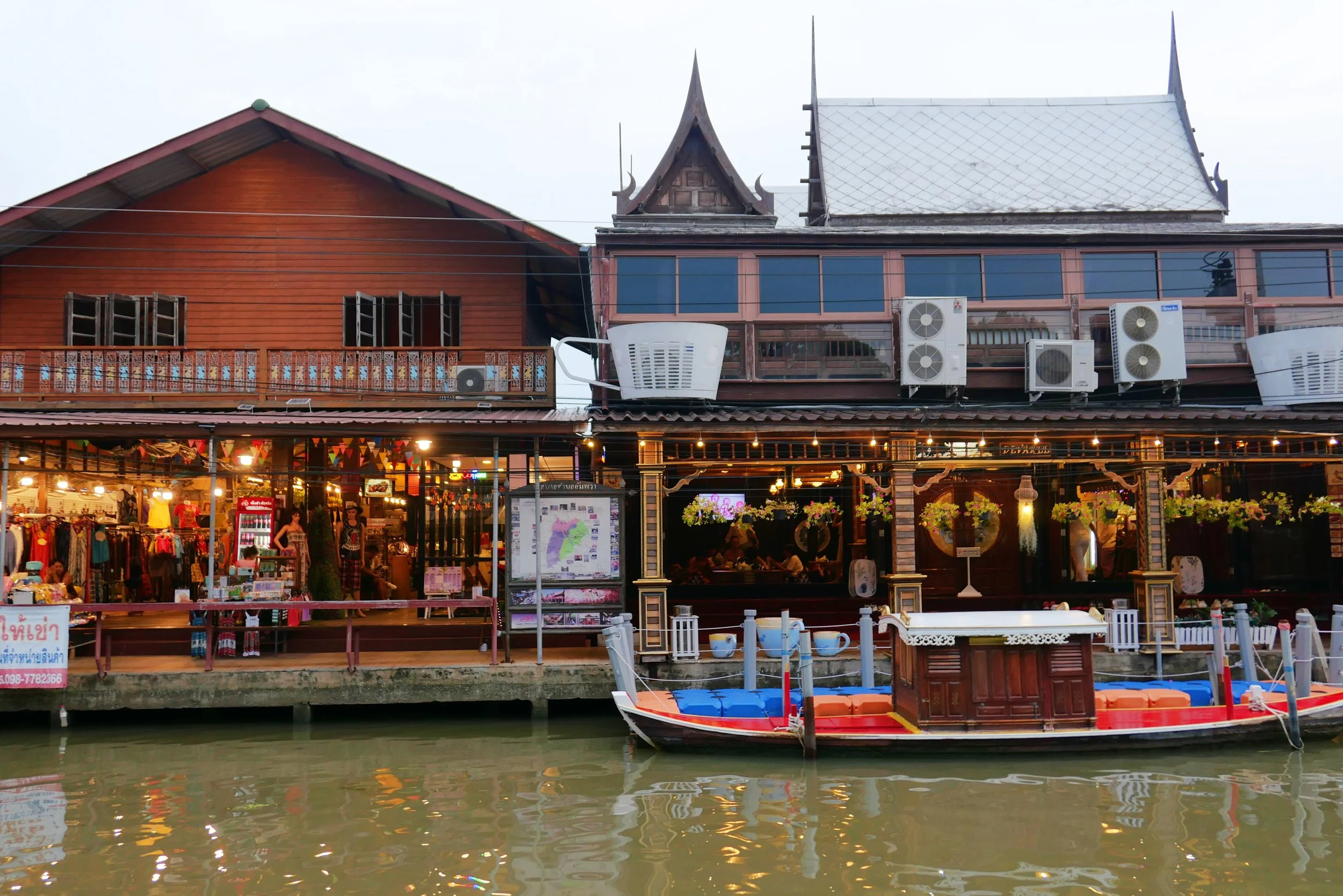 A lively riverside scene with a traditional Thai wooden building and shops, a boat docked at the water's edge, and string lights hanging above, at dusk.