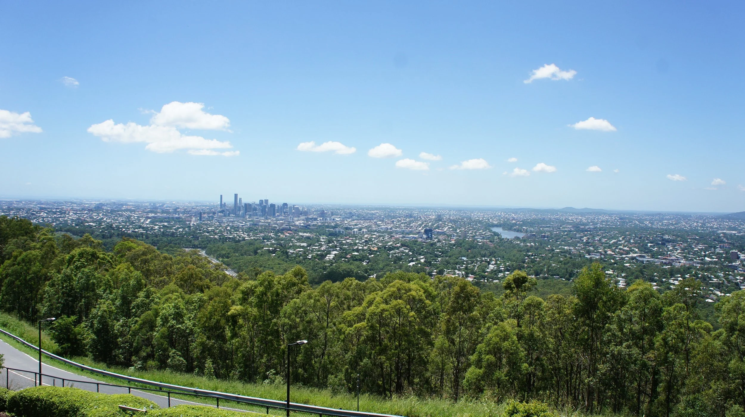 A panoramic view of a city skyline with tall buildings in the distance, surrounded by green trees and a river, under a blue sky with scattered clouds.