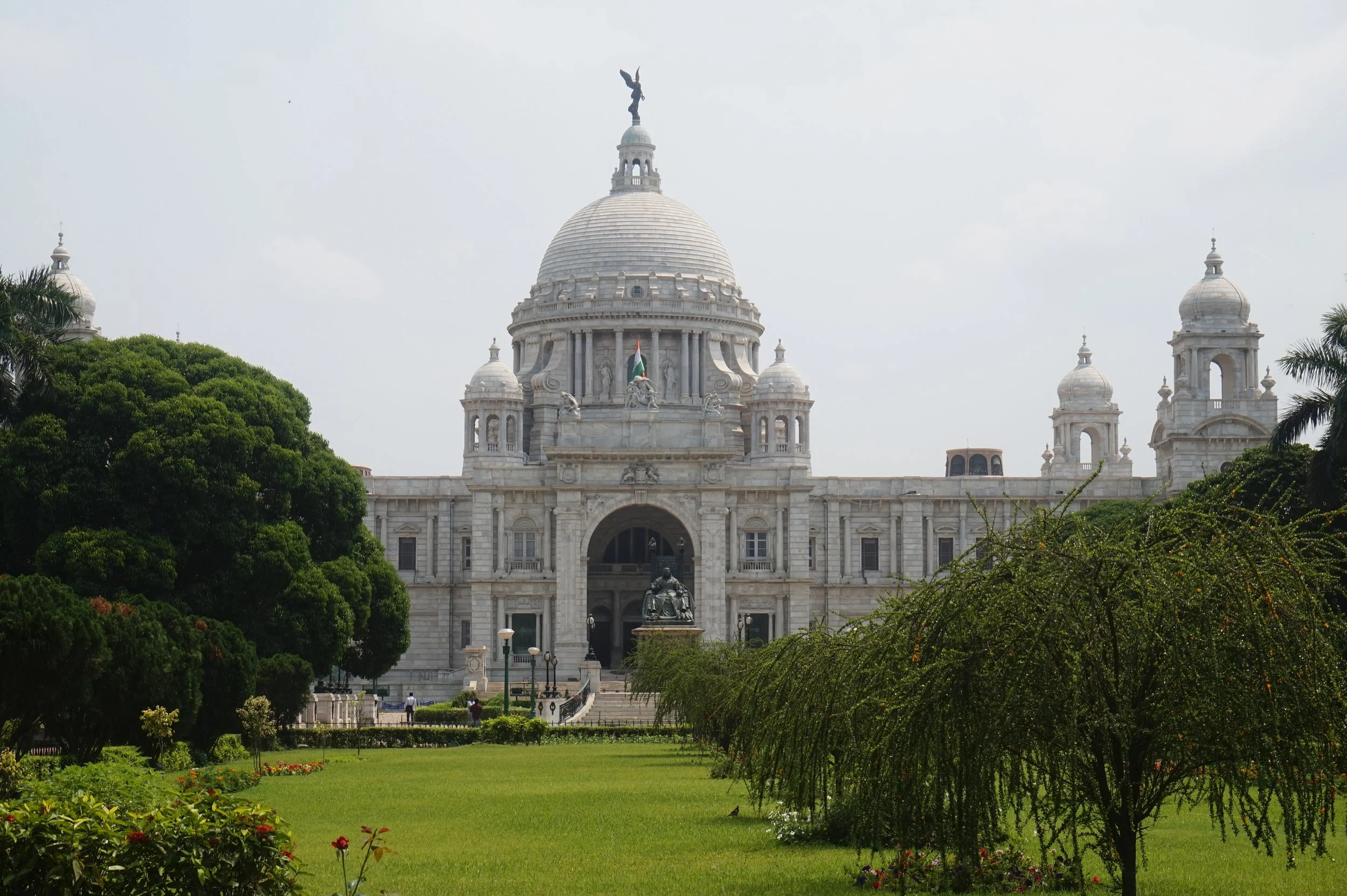 A grand white marble building with a large central dome topped with a statue of an angel holding a wreath, surrounded by smaller domes and columns, set against a cloudy sky, with lush green trees and a manicured lawn in the foreground.