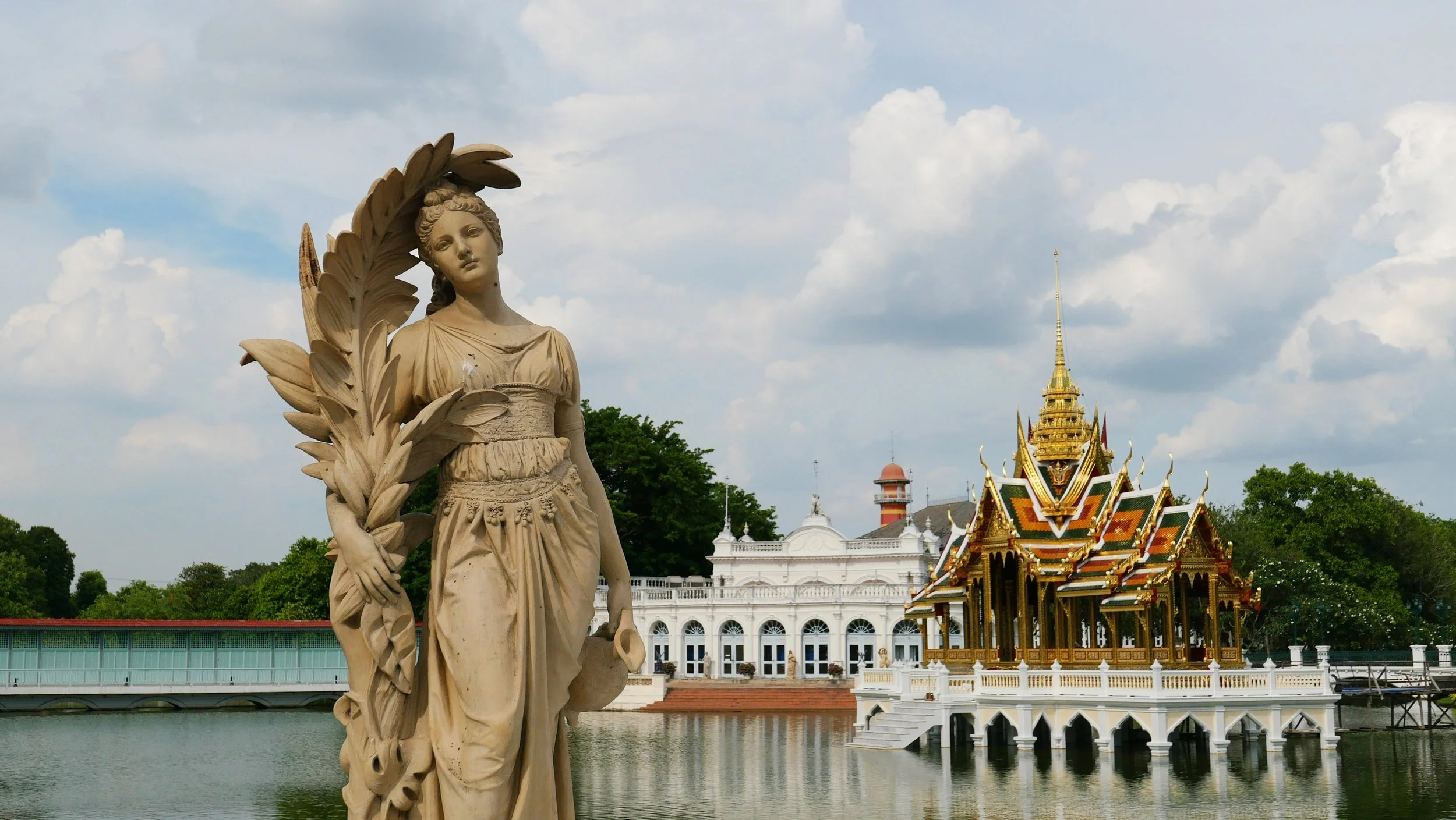 A cream-colored statue of a woman draped in classical attire, holding a staff and surrounded by carved leaves, standing by a pond. In the background, there is a traditional Thai temple with ornate, colorful roofing and a golden spire, along with hist