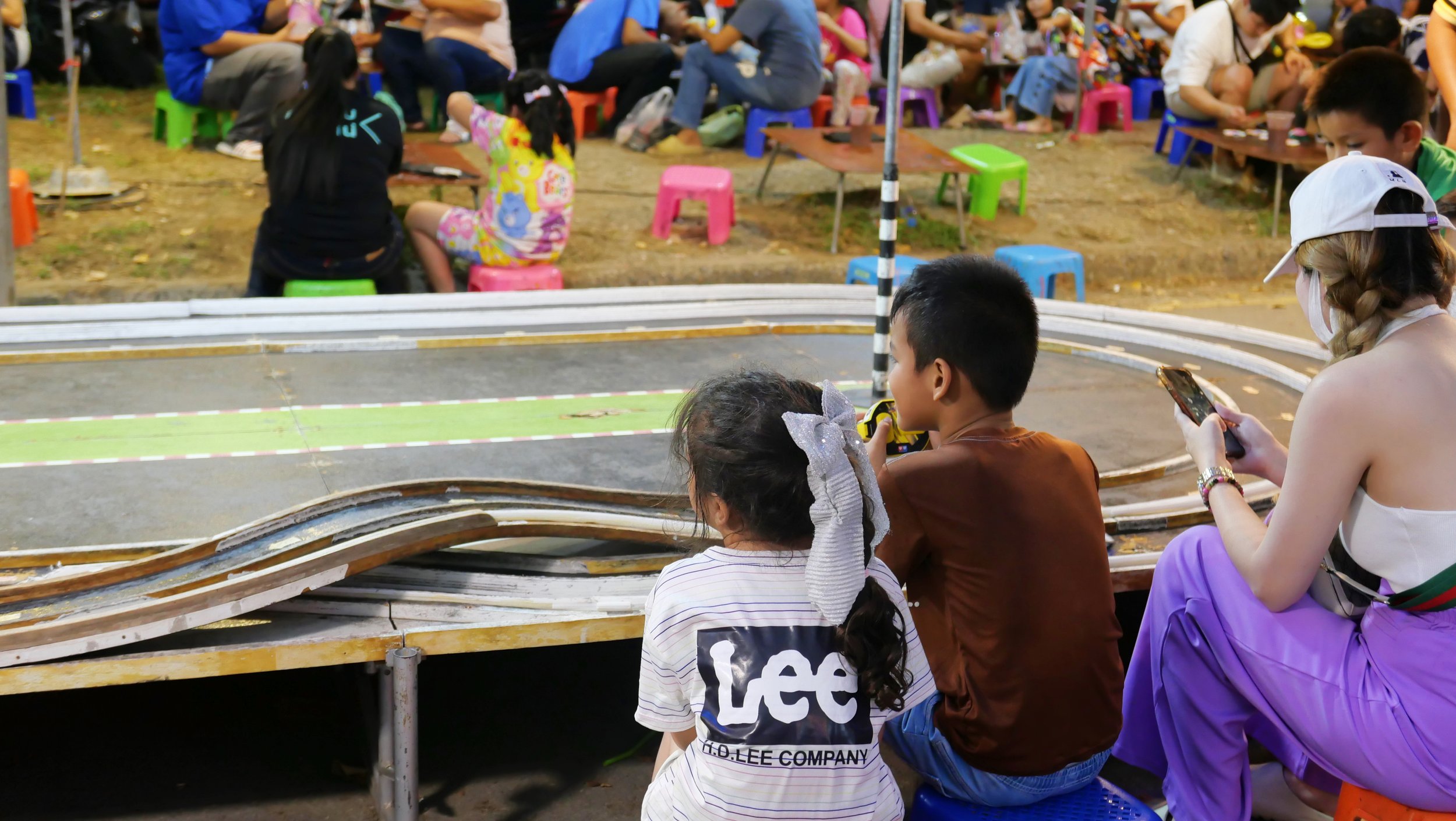 Children and adults gathered outdoors around a model race track, with some children sitting and watching the race and others playing or using devices.