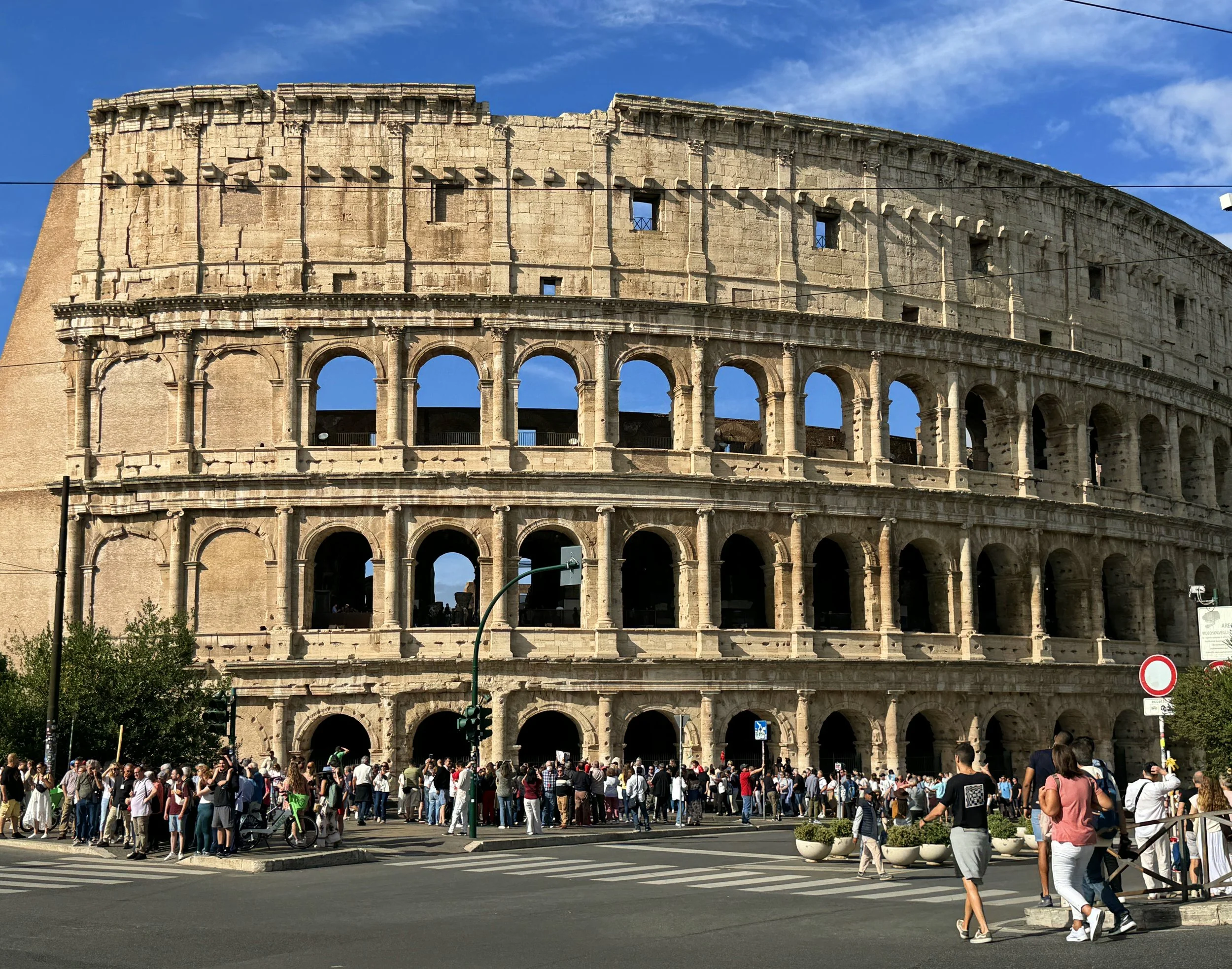 The Roman Colosseum with a crowd of tourists gathered in front of it on a sunny day.
