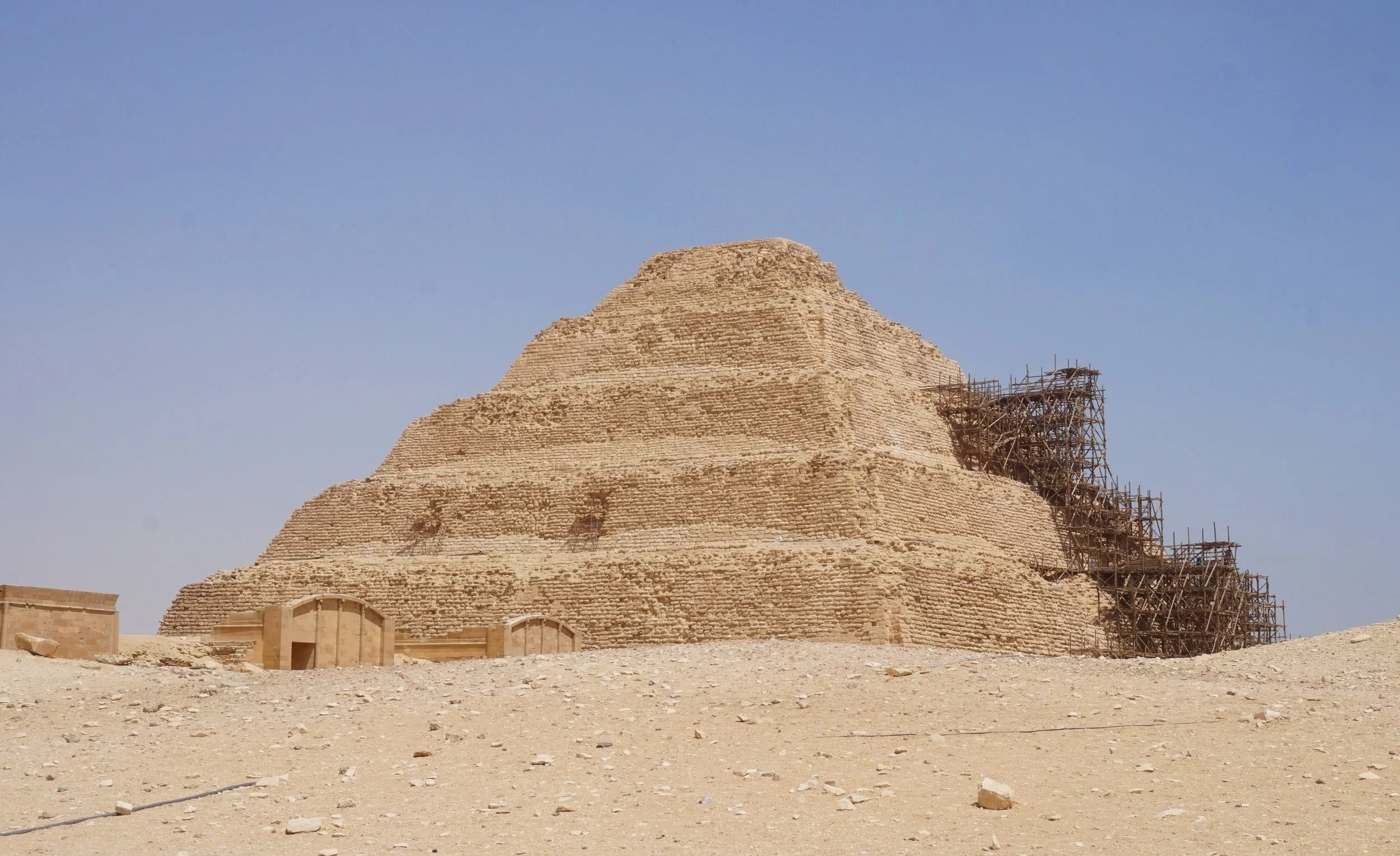 The step pyramid of Djoser, an ancient Egyptian step pyramid under restoration with scaffolding on the right side, against a clear blue sky.