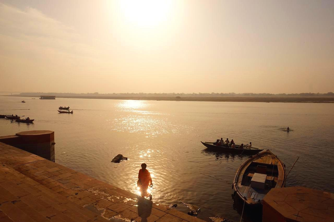 A peaceful river scene at sunset with several boats, including a boat with people, floating on the water. A person is standing on the riverbank near the water's edge, and the scene is bathed in warm, golden sunlight.