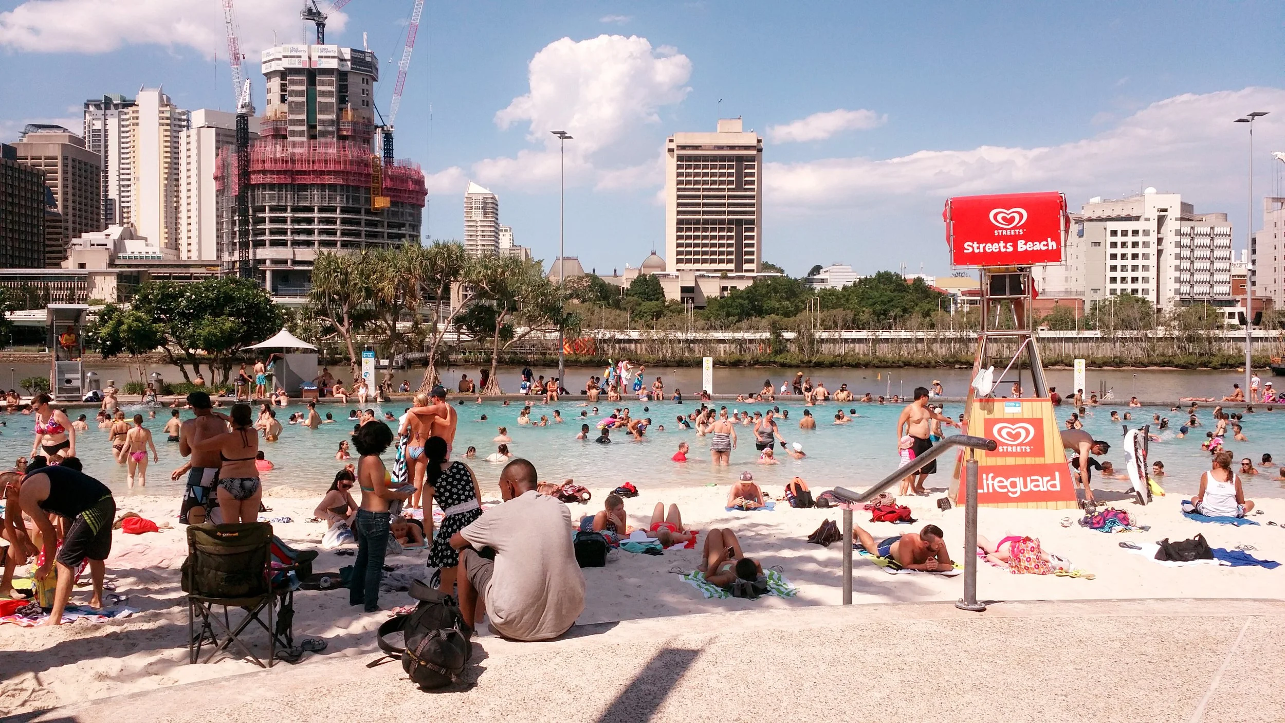 Crowded urban beach scene with many people swimming and relaxing in a pool and on the sand, high-rise buildings in the background, a red Lifeguard tower, and a sign that reads 'Streets Beach' and 'Life guard'.