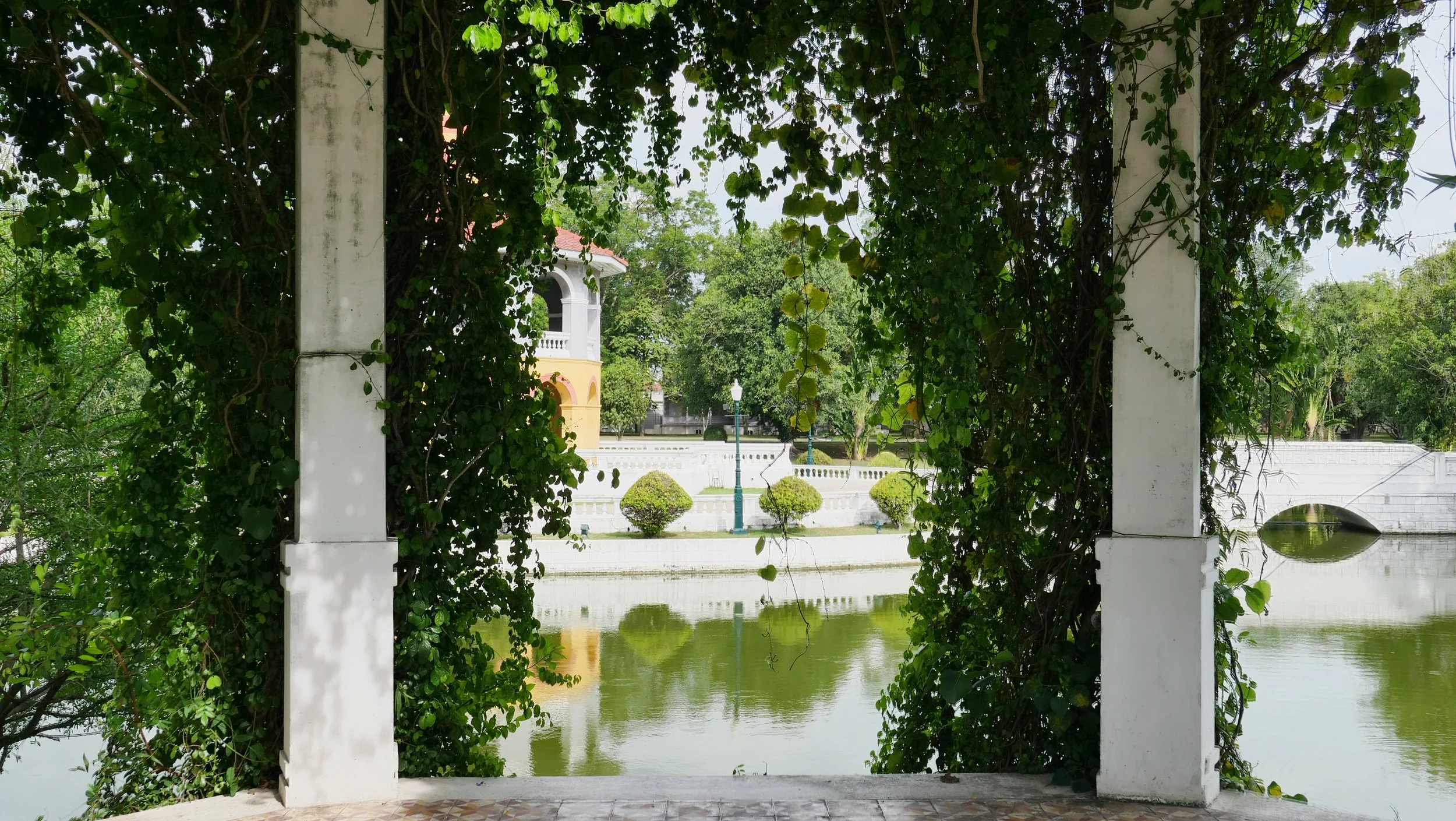 A view of a park or garden area framed by a white arch covered in green vines, overlooking a pond with water toys, neatly trimmed shrubs, a small building with a red roof, and surrounding trees.