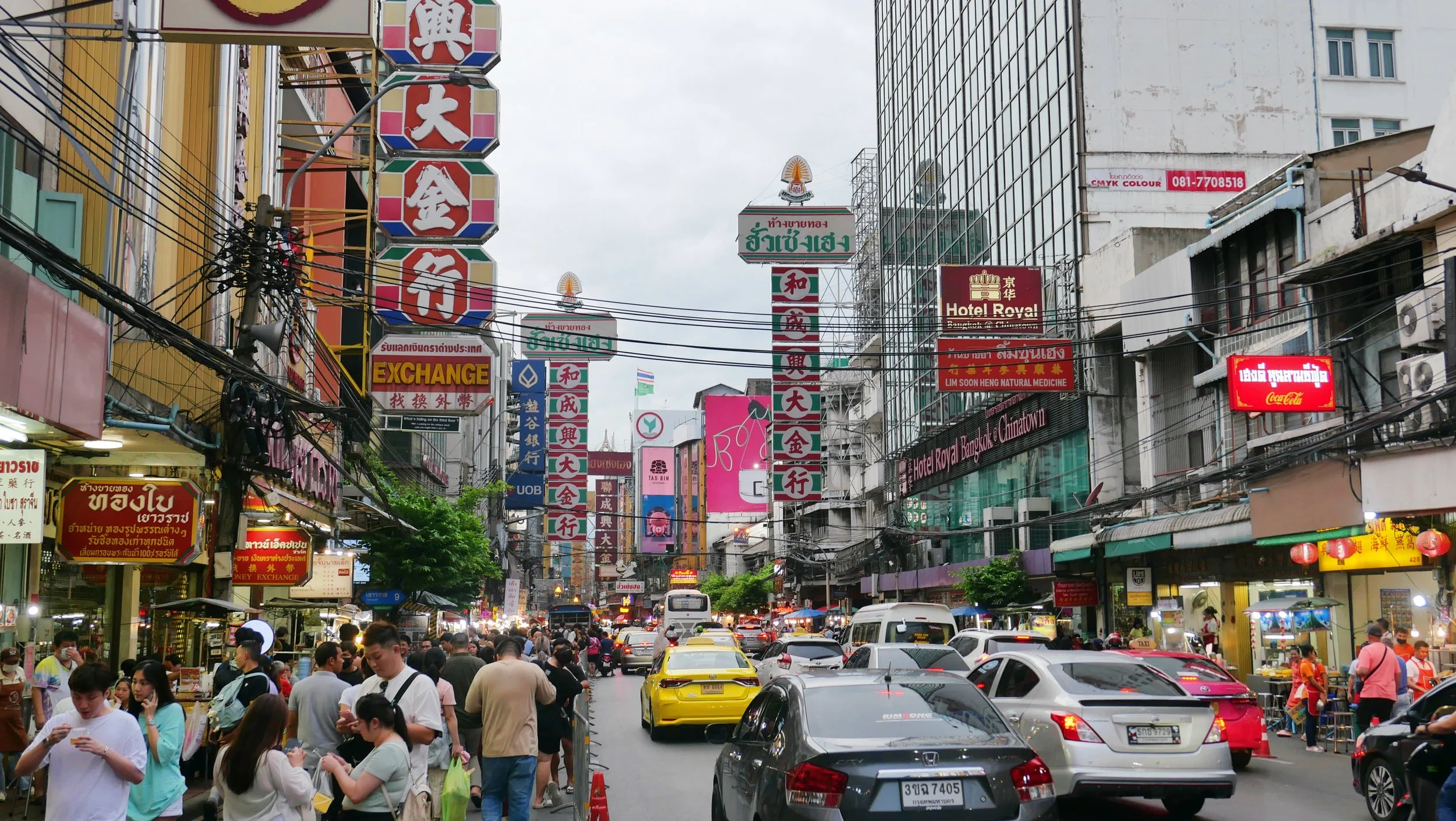 A busy urban street scene in Bangkok with many colorful signs and advertisements in various languages, including Thai and Chinese. The street is crowded with pedestrians and vehicles, including taxis, and is lined with buildings showing signs for hot