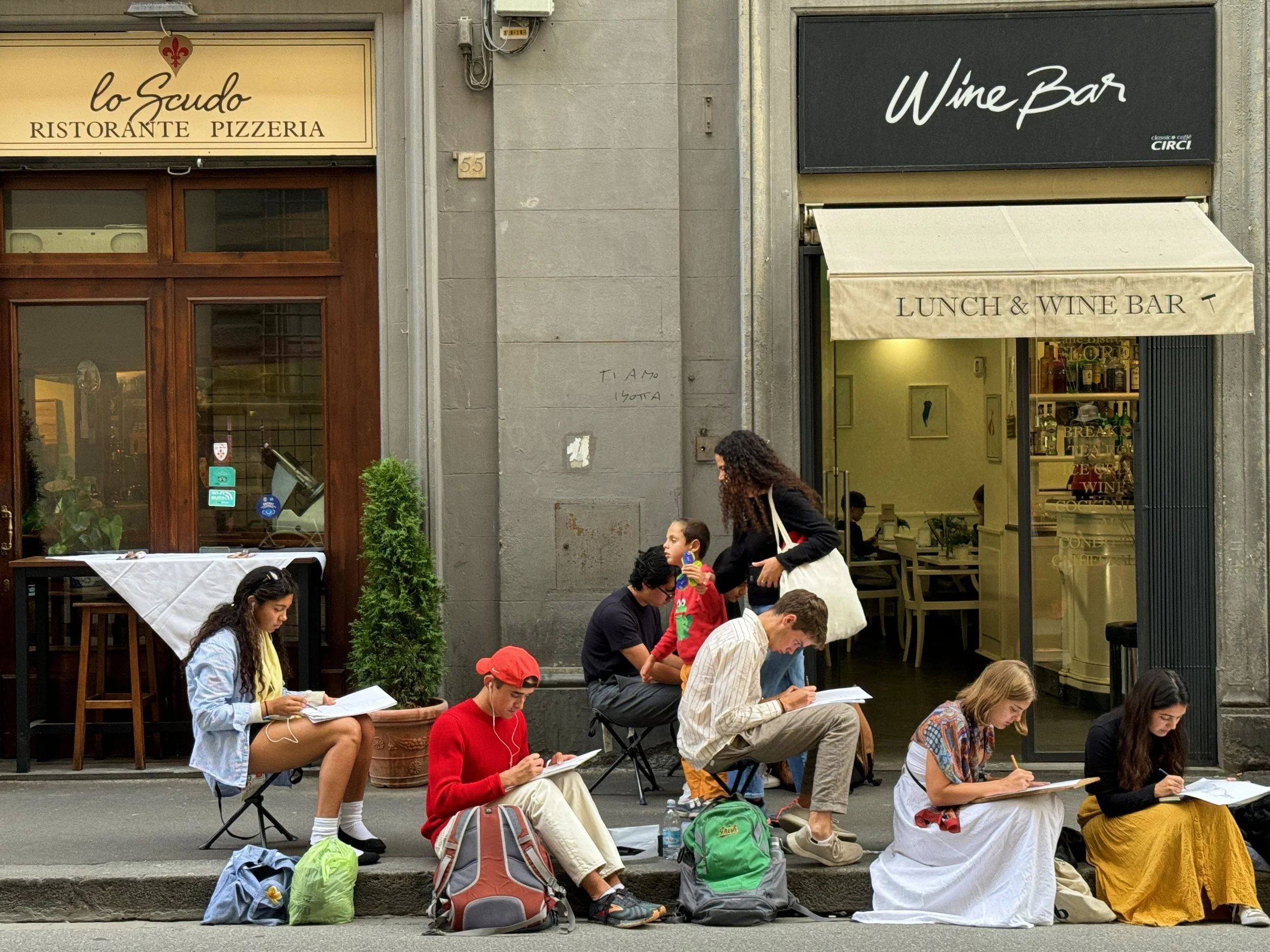 People sitting and standing outside a restaurant and wine bar, taking notes or reading, with backpacks and personal items around them.